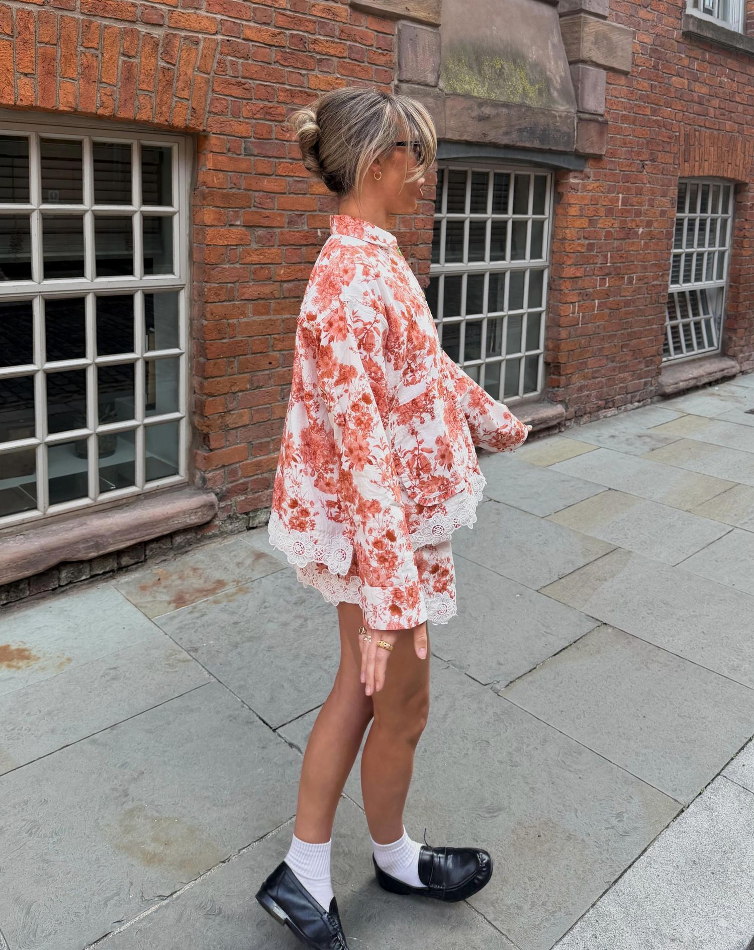 Woman in floral orange mini dress with lace trim and black loafers poses on stone pavement outside brick building.