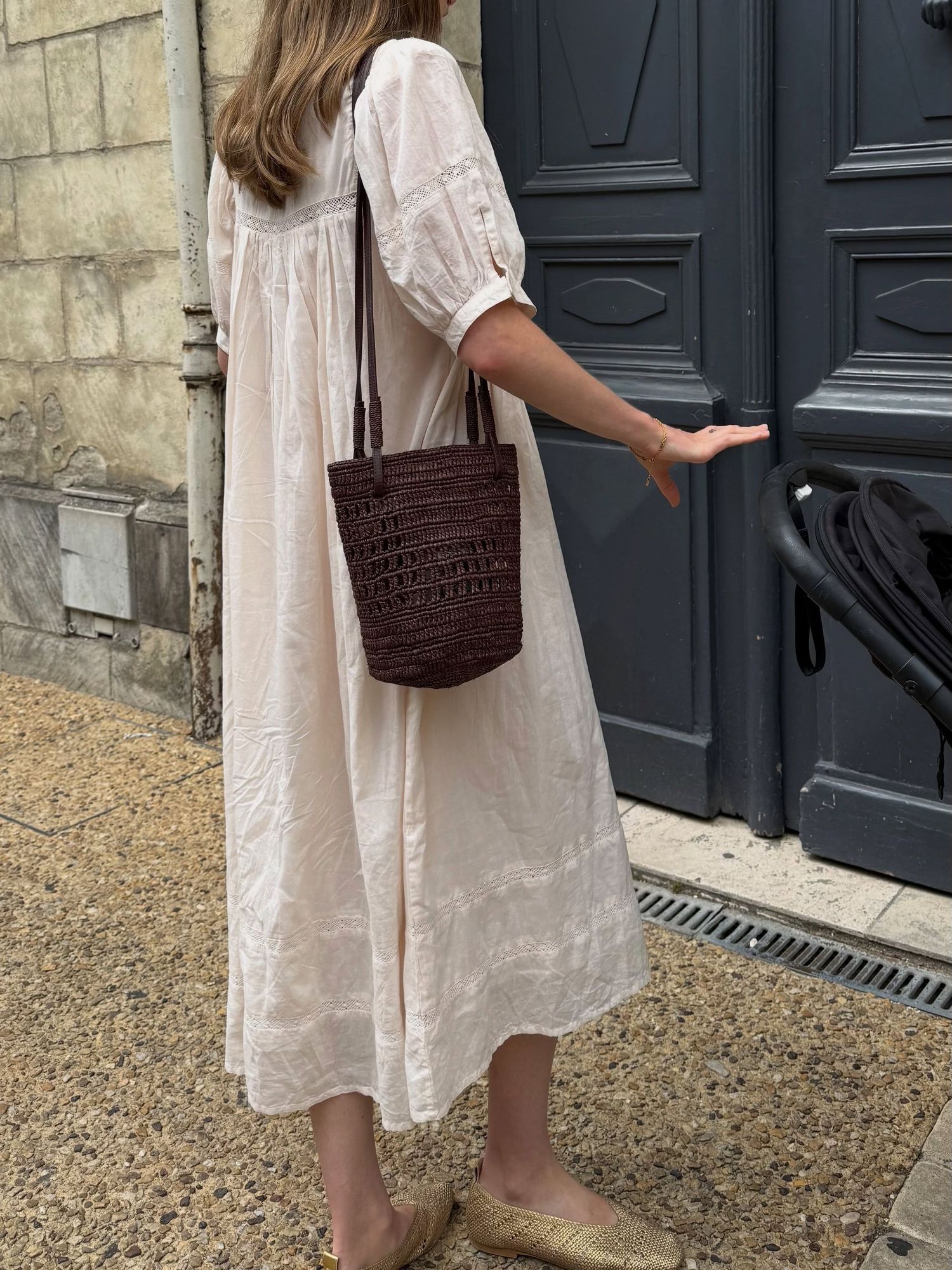 Woman in white linen midi dress with puff sleeves, carrying dark brown woven bucket bag, wearing beige mules.