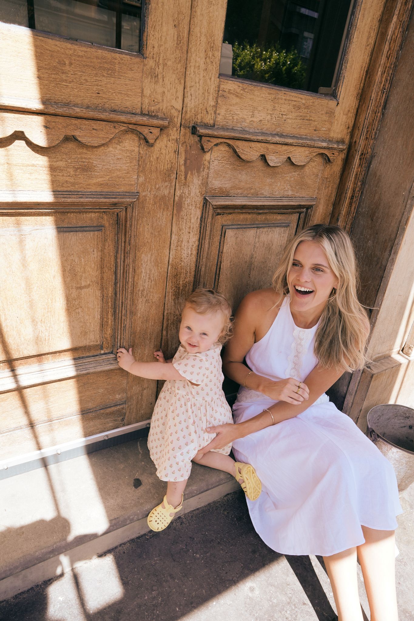 Woman in white dress laughing with baby girl in floral outfit on wooden door steps.