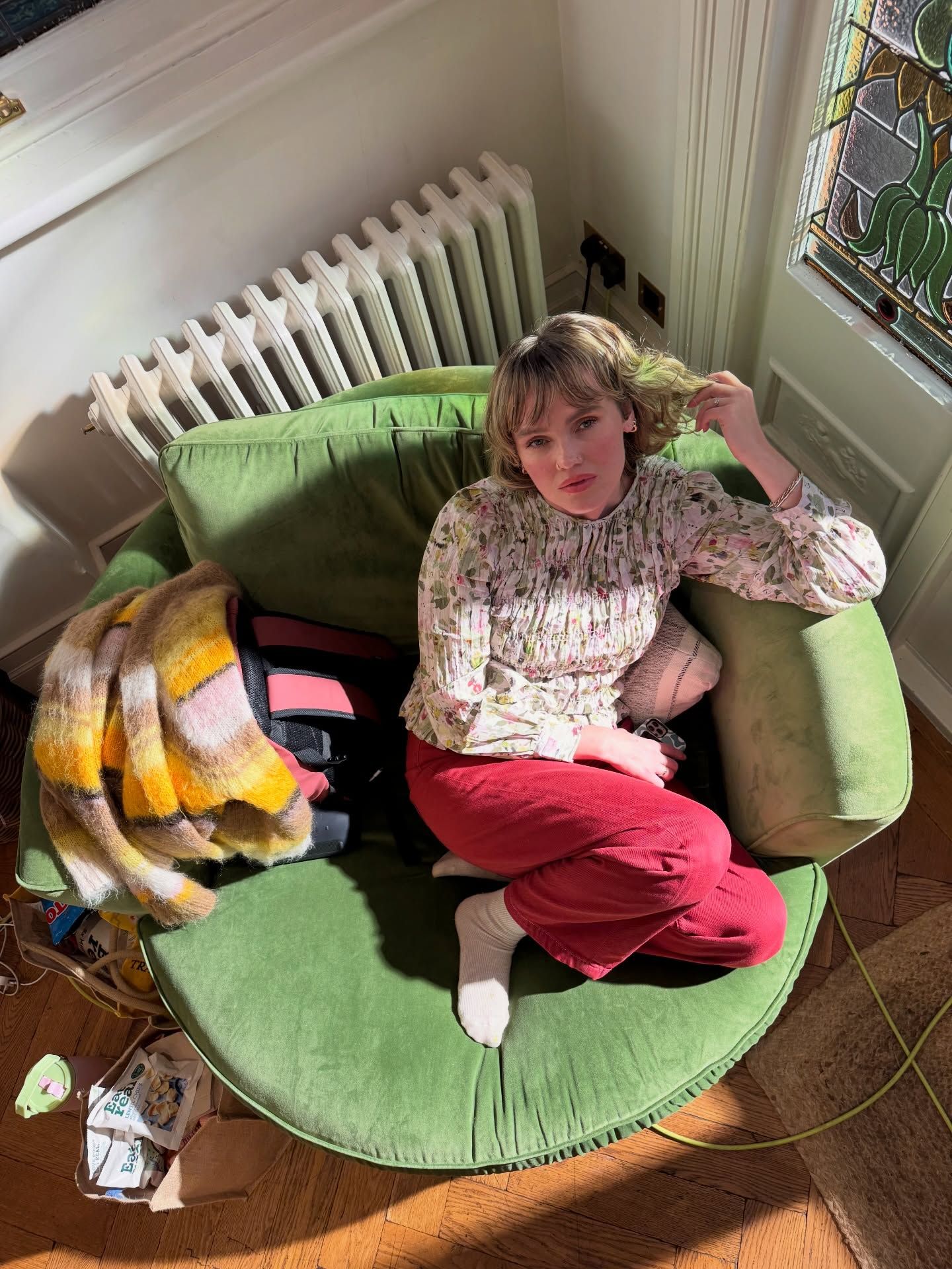 Young woman in floral top and red pants sits curled up in a green armchair by a stained glass window.