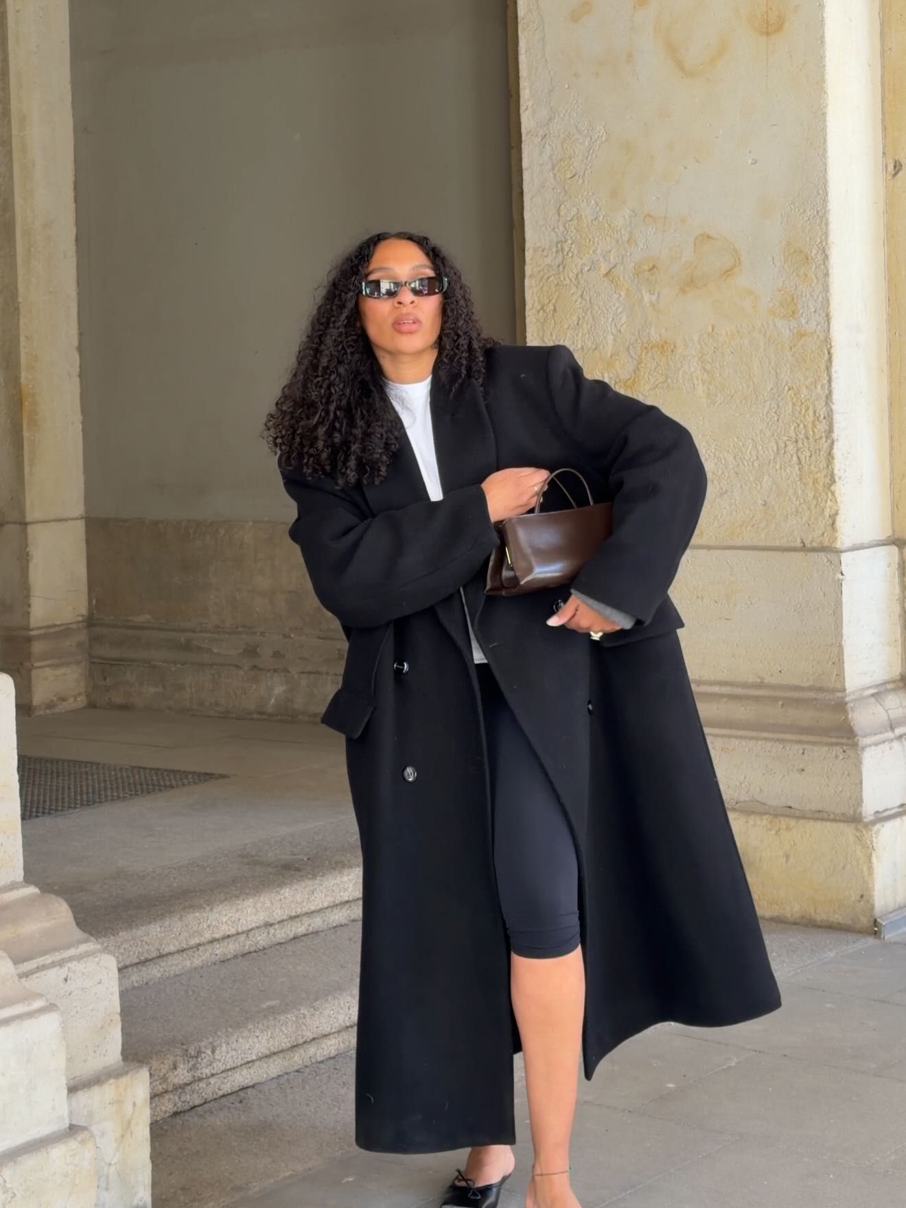 Woman in oversized black coat, bike shorts, sunglasses holding brown bag outside stone building.