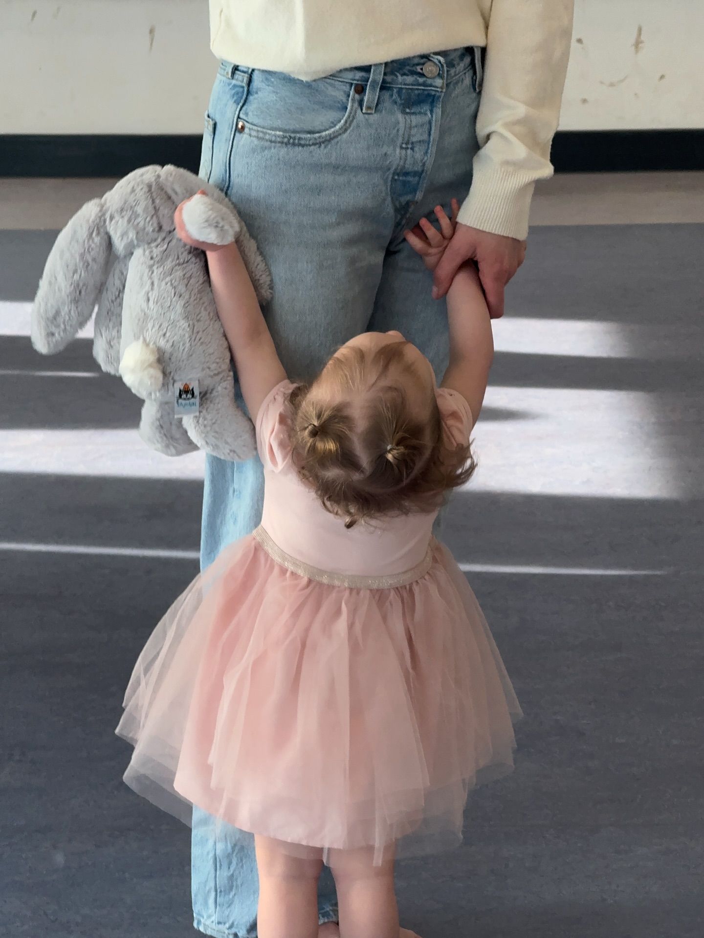 Toddler girl in pink tutu dress holds stuffed bunny while clinging to adult in jeans legs from behind.