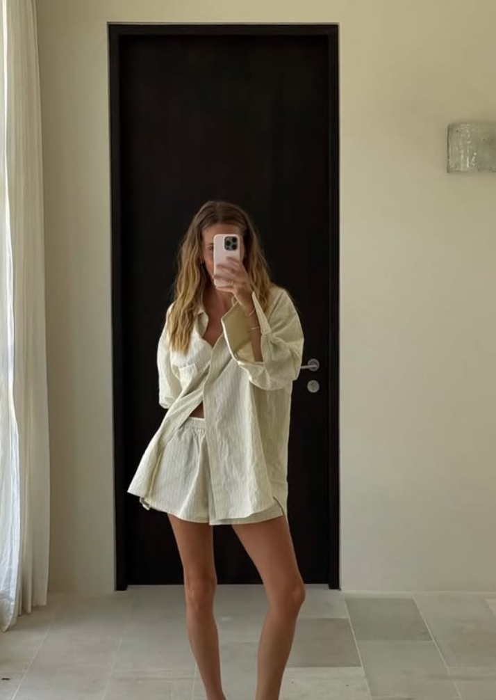 Person in white oversized shirt poses in front of mirror while taking selfies with smartphone.