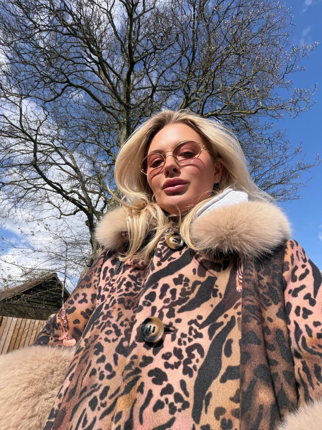 Blonde woman in leopard print fur-collar coat and pink sunglasses outdoors on a sunny day.