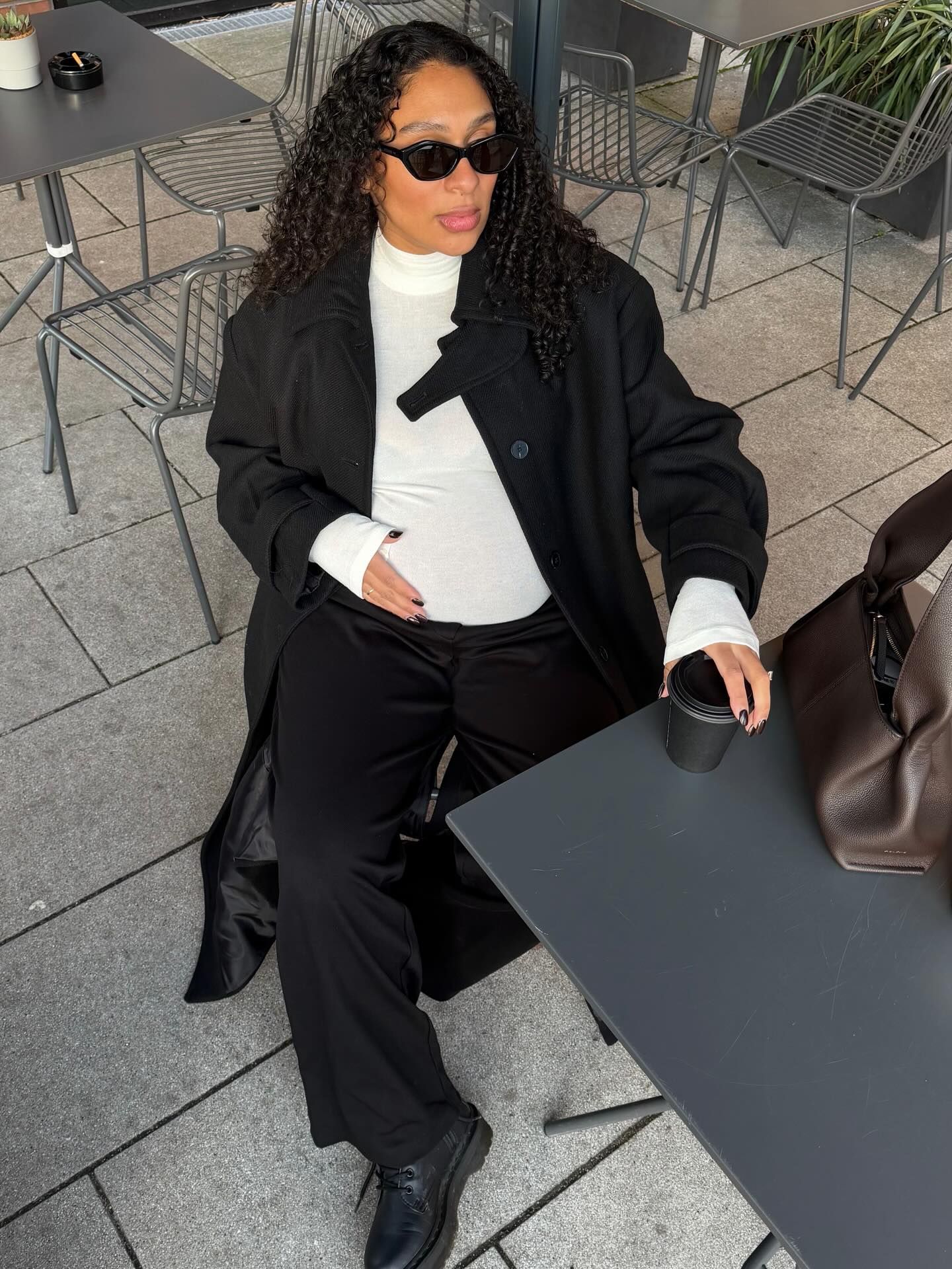Woman in black coat, white turtleneck, and black trousers sitting at outdoor cafe table with sunglasses.