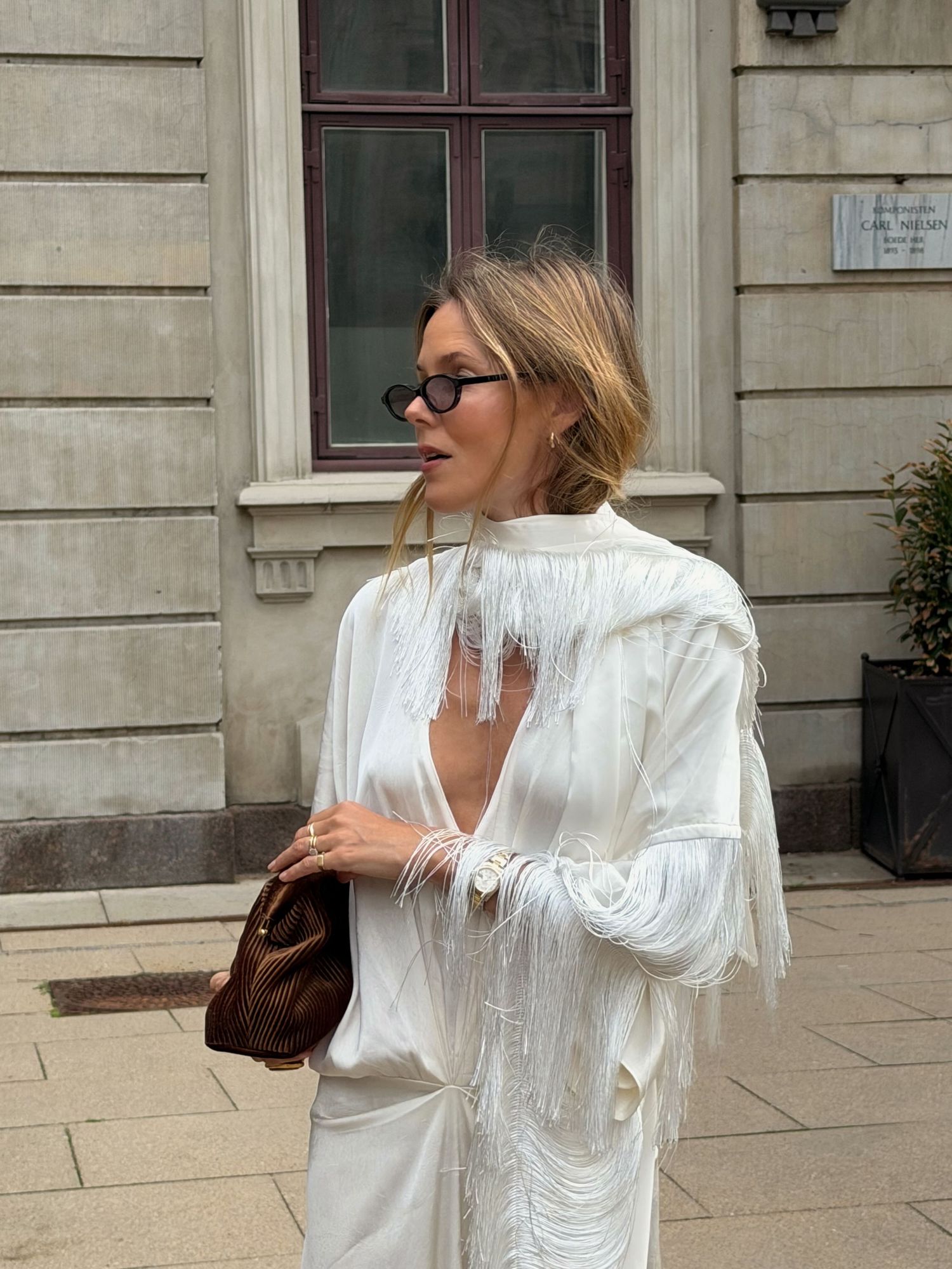 Woman in white fringed dress with sunglasses holding brown bag outside classic European building.