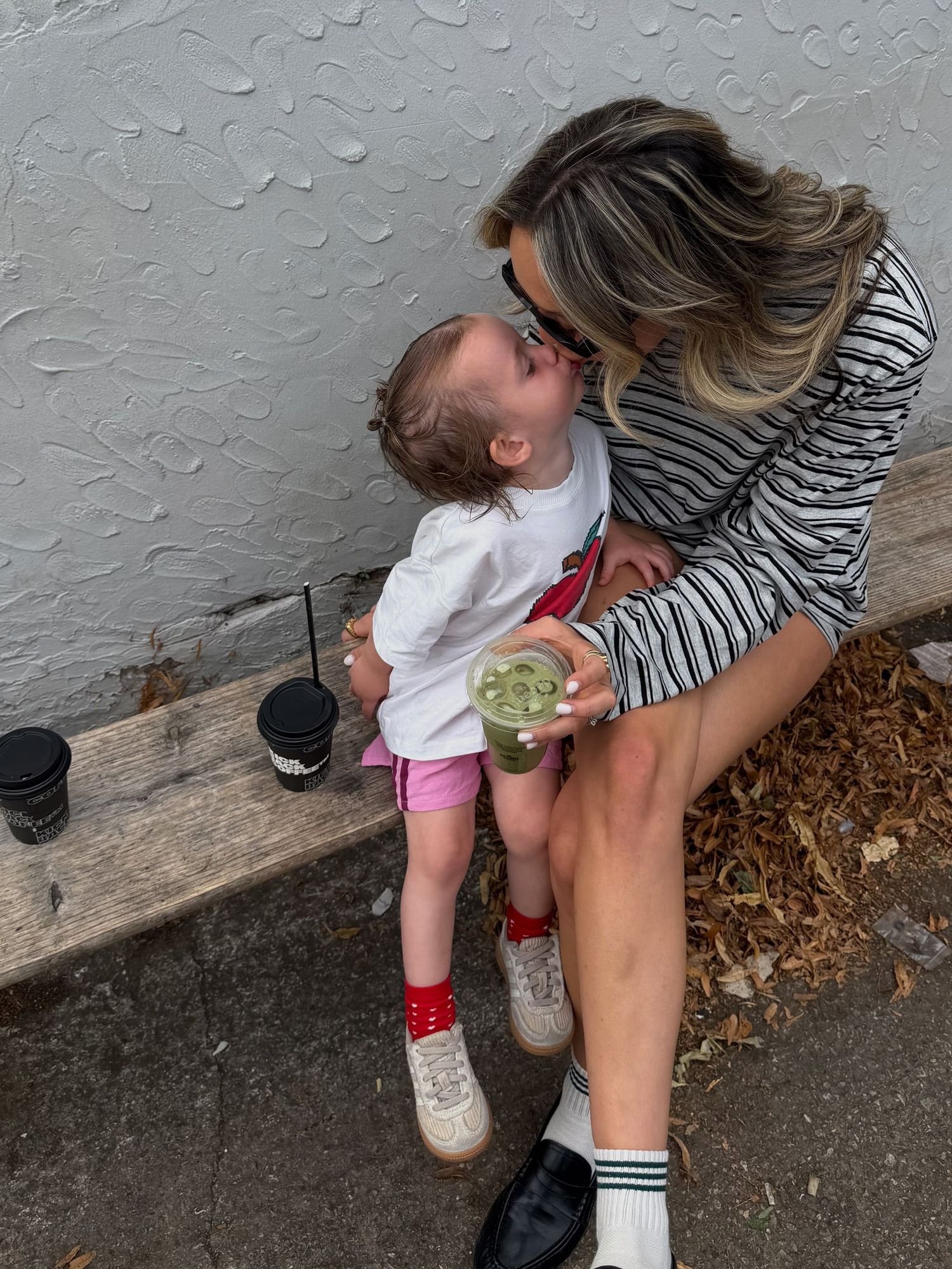 A woman sitting on a bench kisses a toddler girl while holding a green drink outdoors.