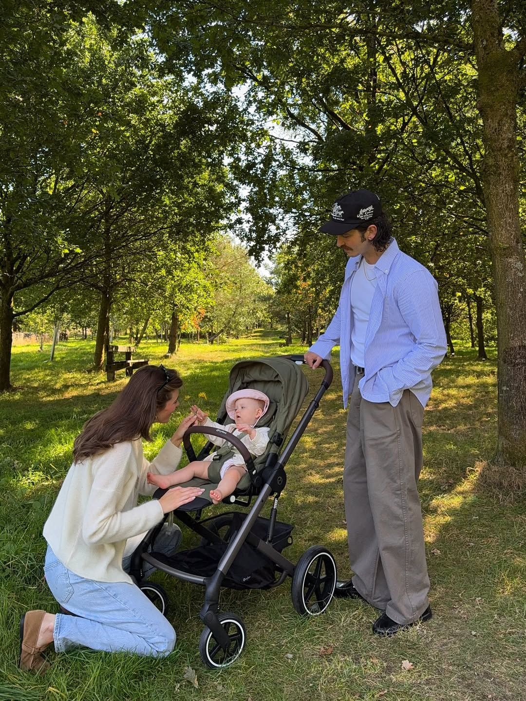 Young family with baby in green stroller enjoying a sunny day in a lush park setting.