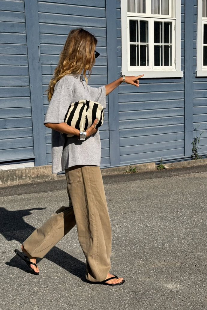 Woman in gray tee, wide-leg linen pants, and sandals holding striped clutch bag, walking past blue building.