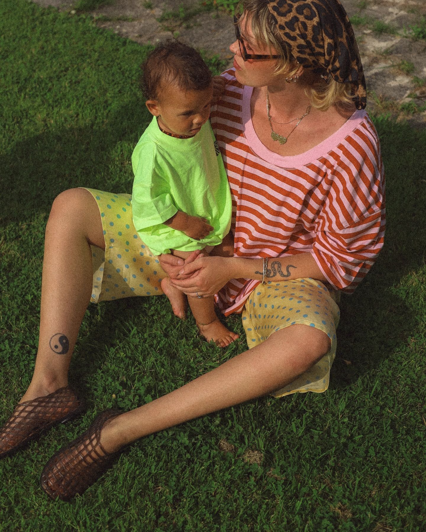 Woman in striped top holding a baby in neon green shirt, sitting on grass wearing yellow polka dot shorts.