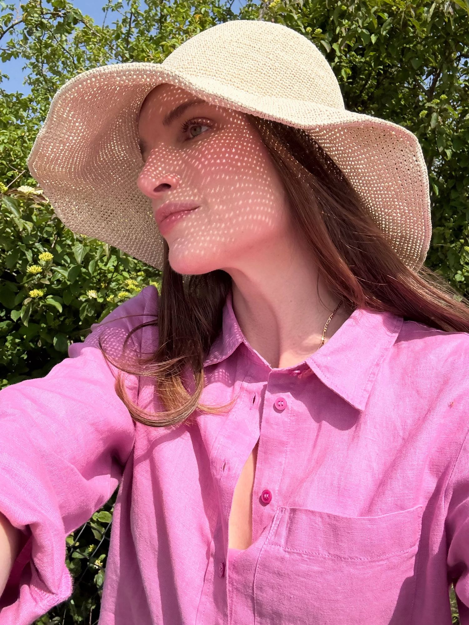 Woman in pink linen shirt and woven sun hat enjoying sunny day outdoors with green foliage background.