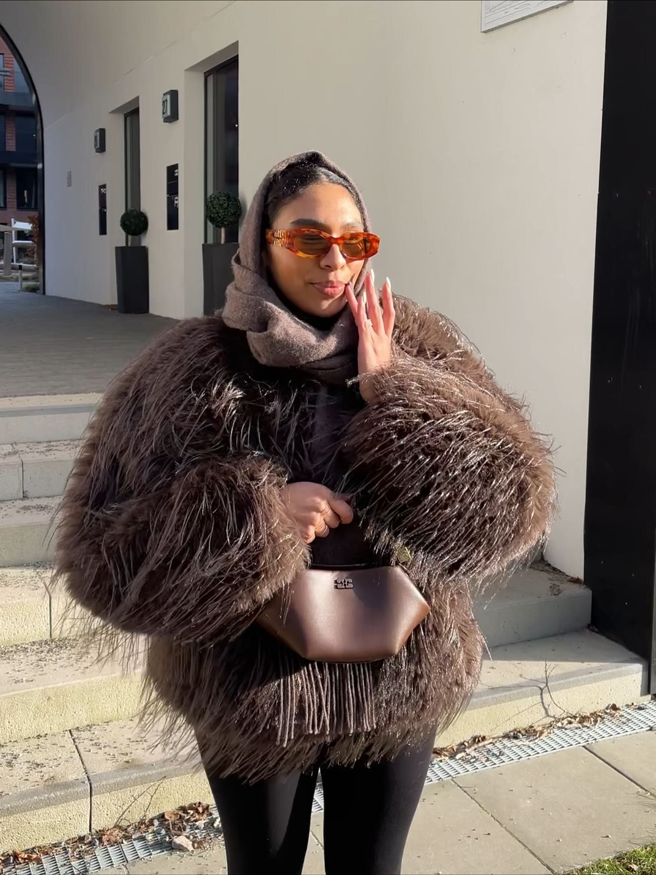Woman in fluffy brown fur coat, orange sunglasses, holding a designer bag, posing outdoors in winter.