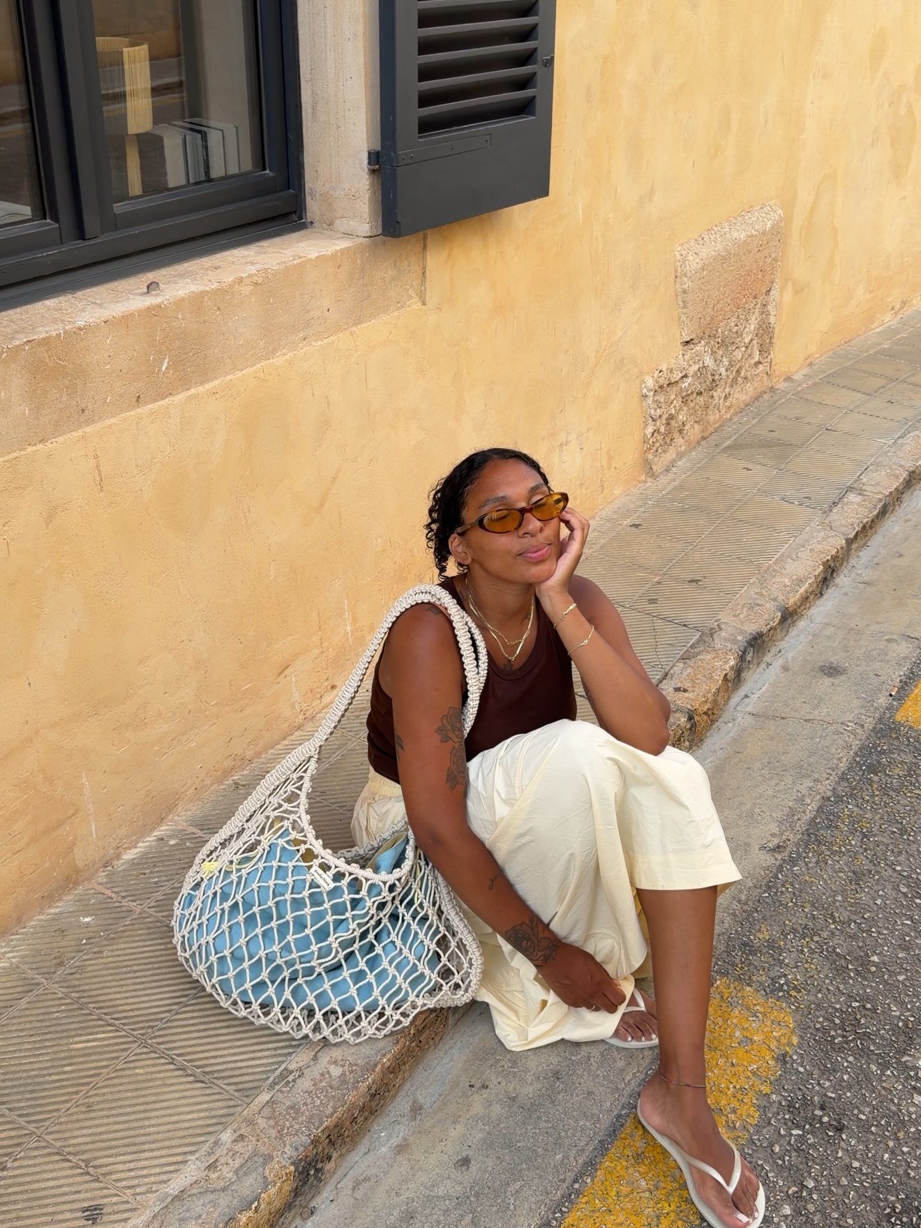 Woman sitting on curb wearing brown top, cream pants, sunglasses, holding a white mesh bag with blue accents.