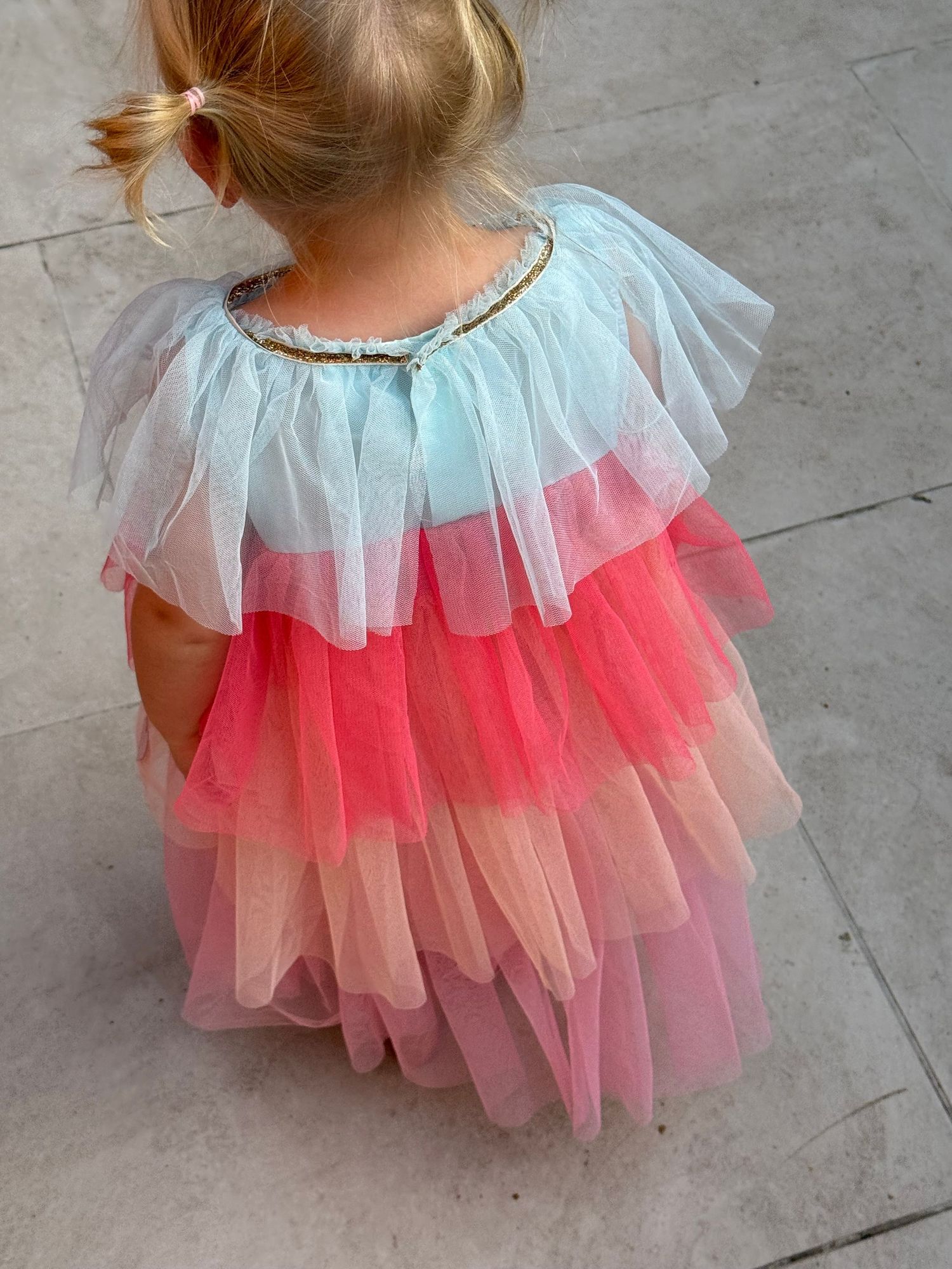 Young girl wearing a tiered rainbow tulle dress in blue, coral, and pink layers with sequin neckline detail.
