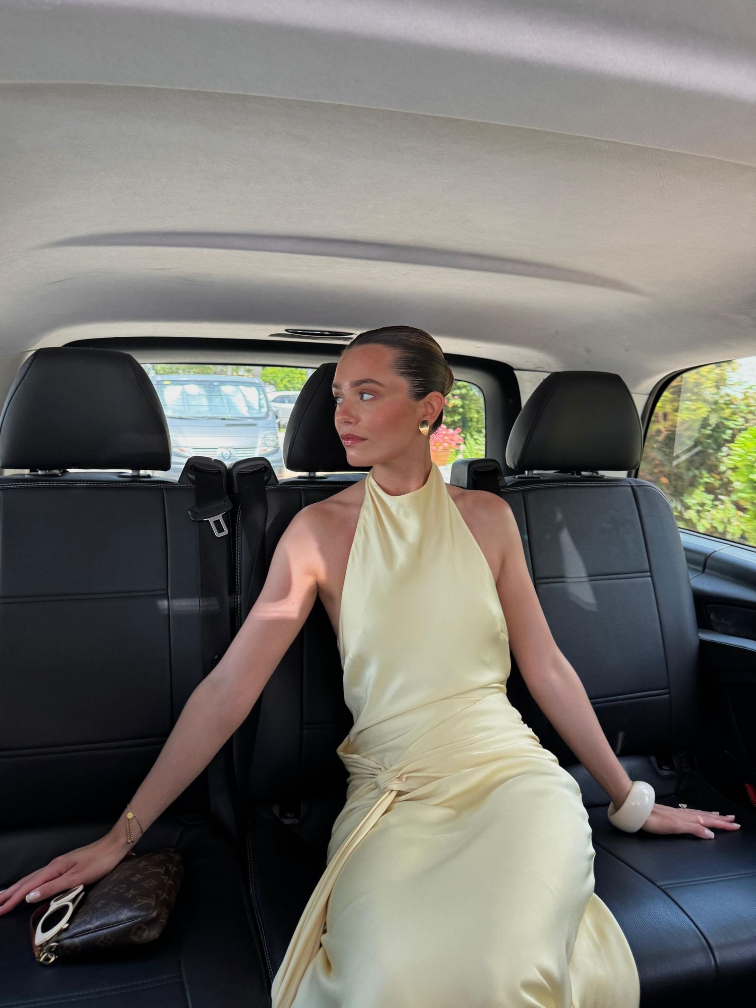 Woman in elegant pale yellow satin halter dress seated in black leather car interior, looking sideways.