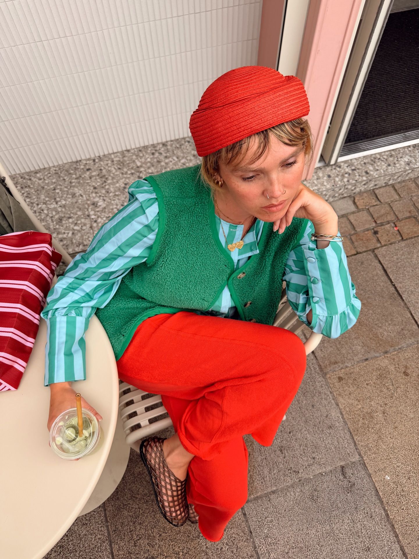 Woman in red hat, green vest, striped shirt, and red pants sits at outdoor cafe table holding a drink.