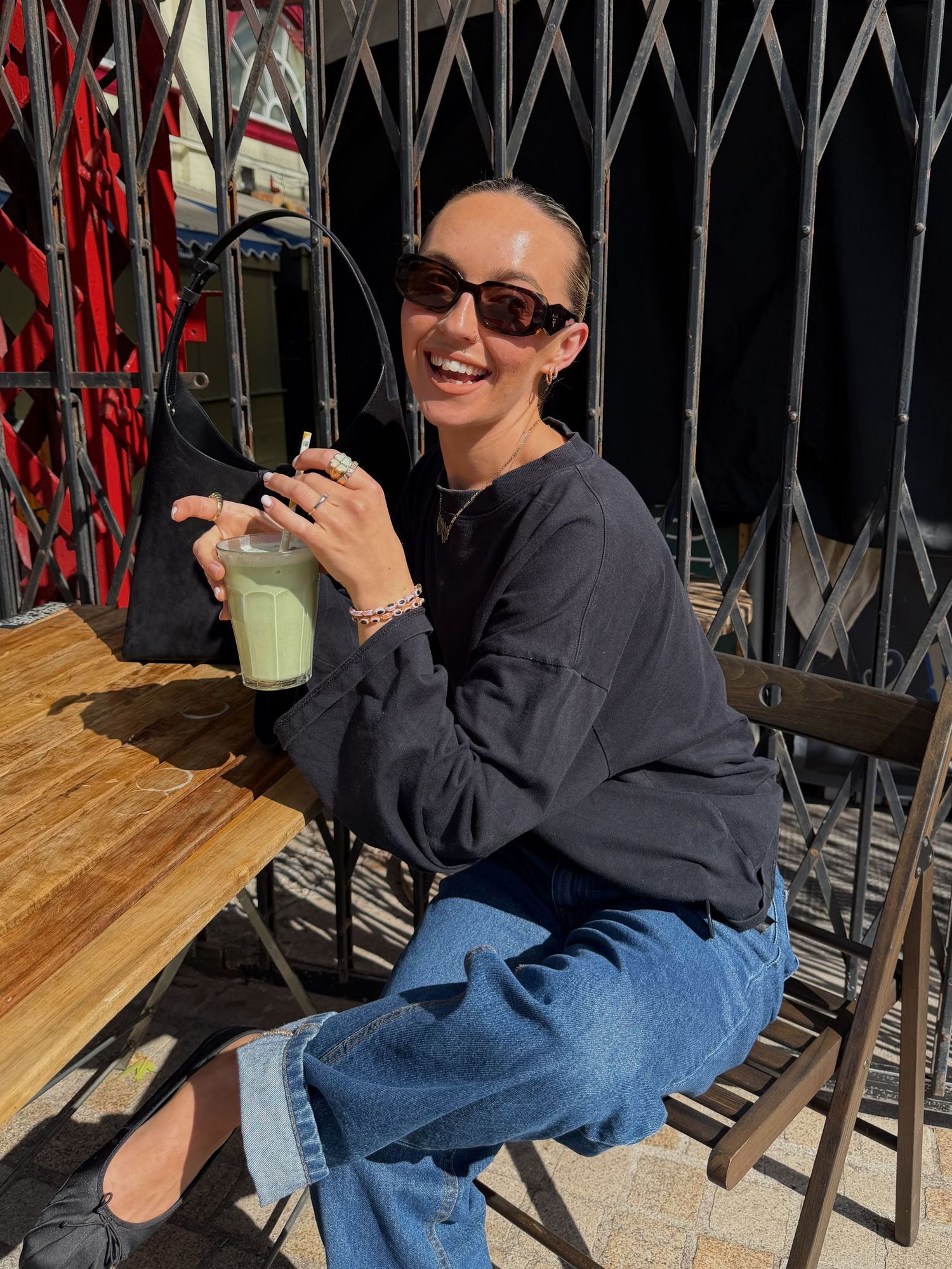 Woman in sunglasses smiling outdoors, wearing dark sweatshirt and jeans, holding green drink at wooden cafe table.