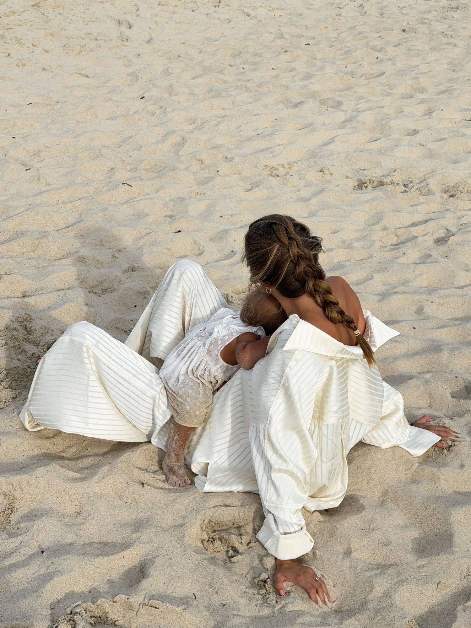 Woman in white striped linen outfit sitting relaxed on sandy beach, showcasing casual boho summer fashion.