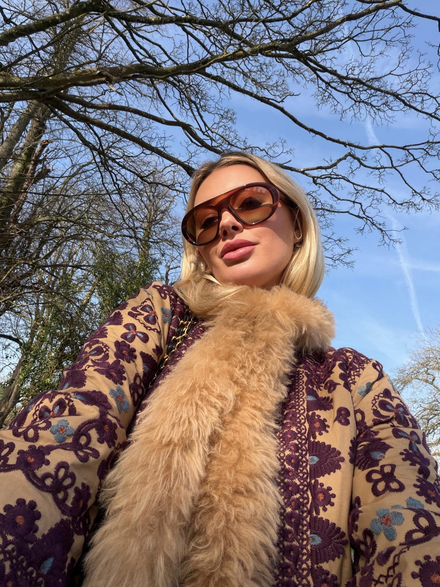 Blonde woman in embroidered coat with fur trim and brown sunglasses posing outdoors under bare trees.