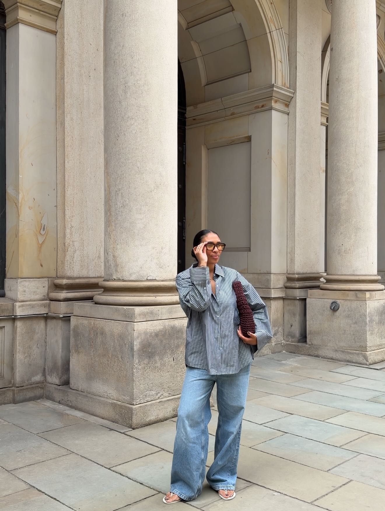 Woman in blue striped shirt and wide-leg jeans poses before grand stone columns of a classical building.
