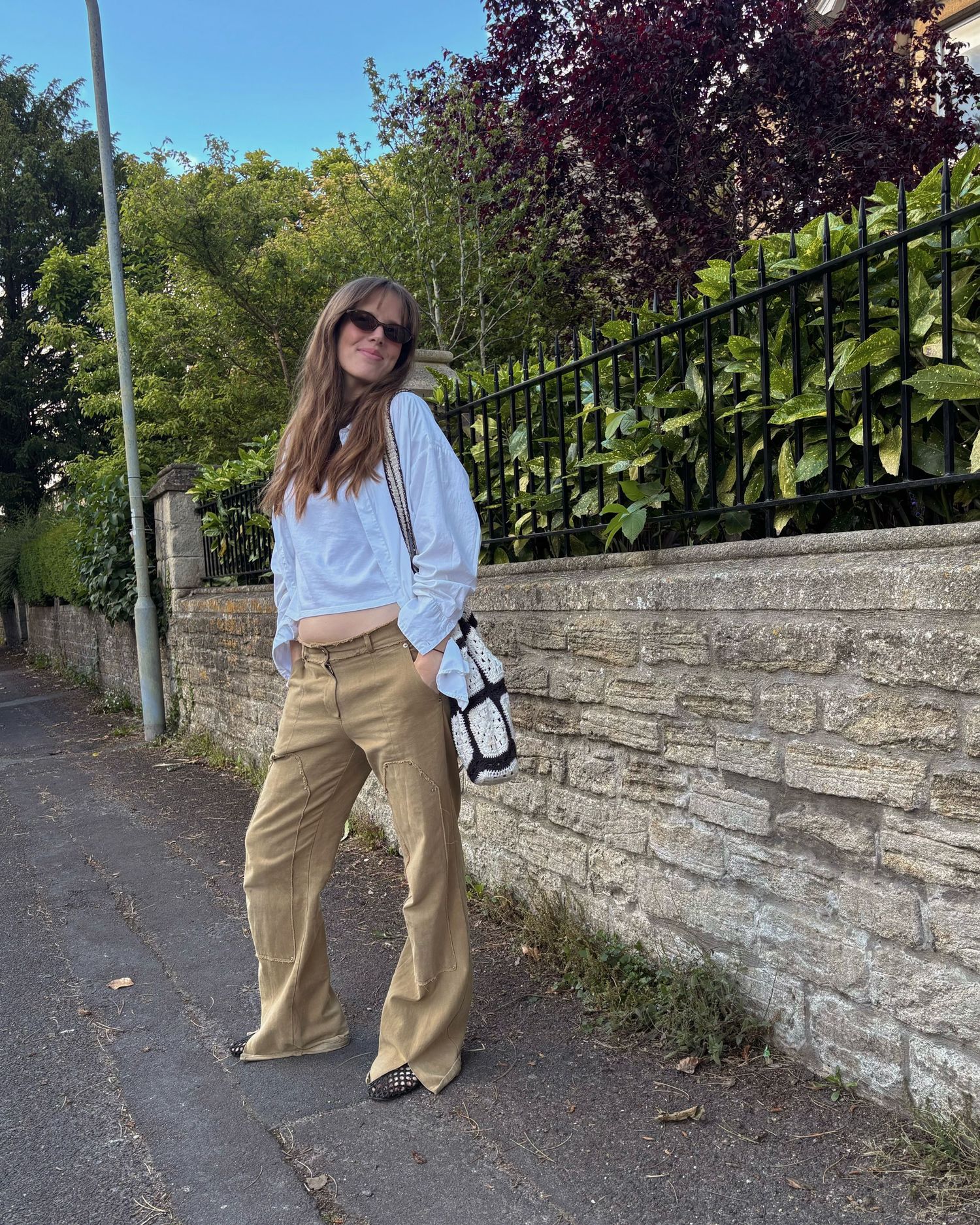 Woman in white cropped shirt, tan wide-leg trousers, sunglasses, and small bag posing on a sunny street.