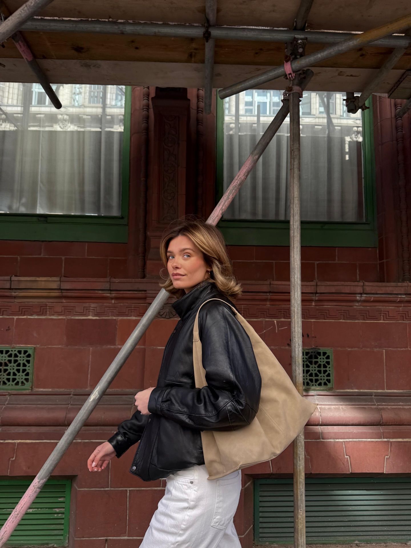 Woman in black leather jacket carrying a large beige suede shoulder bag, posing outdoors near a red brick building.
