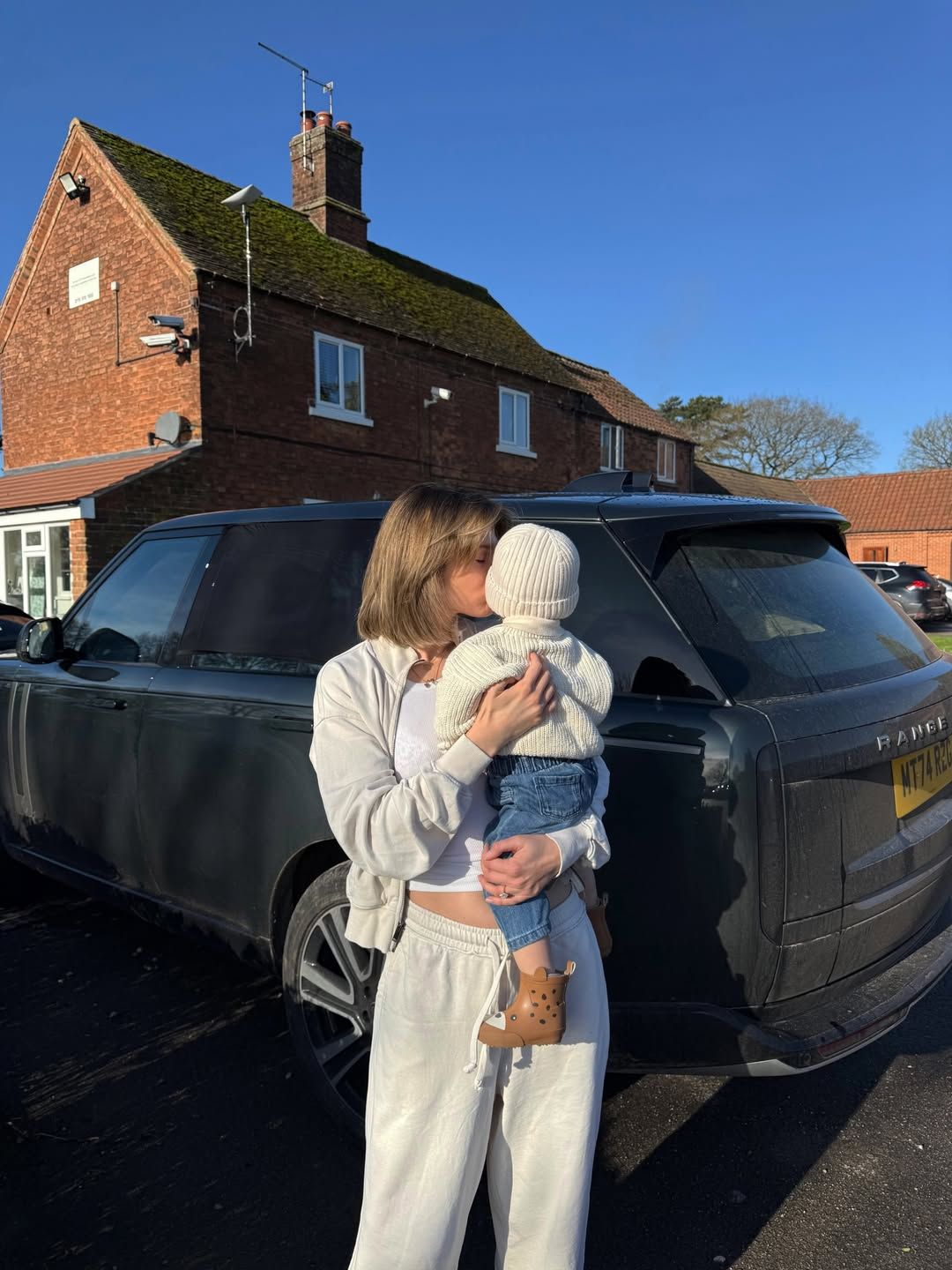 Woman in white outfit kissing a baby in a white hat, standing beside a black Range Rover outside a brick house.