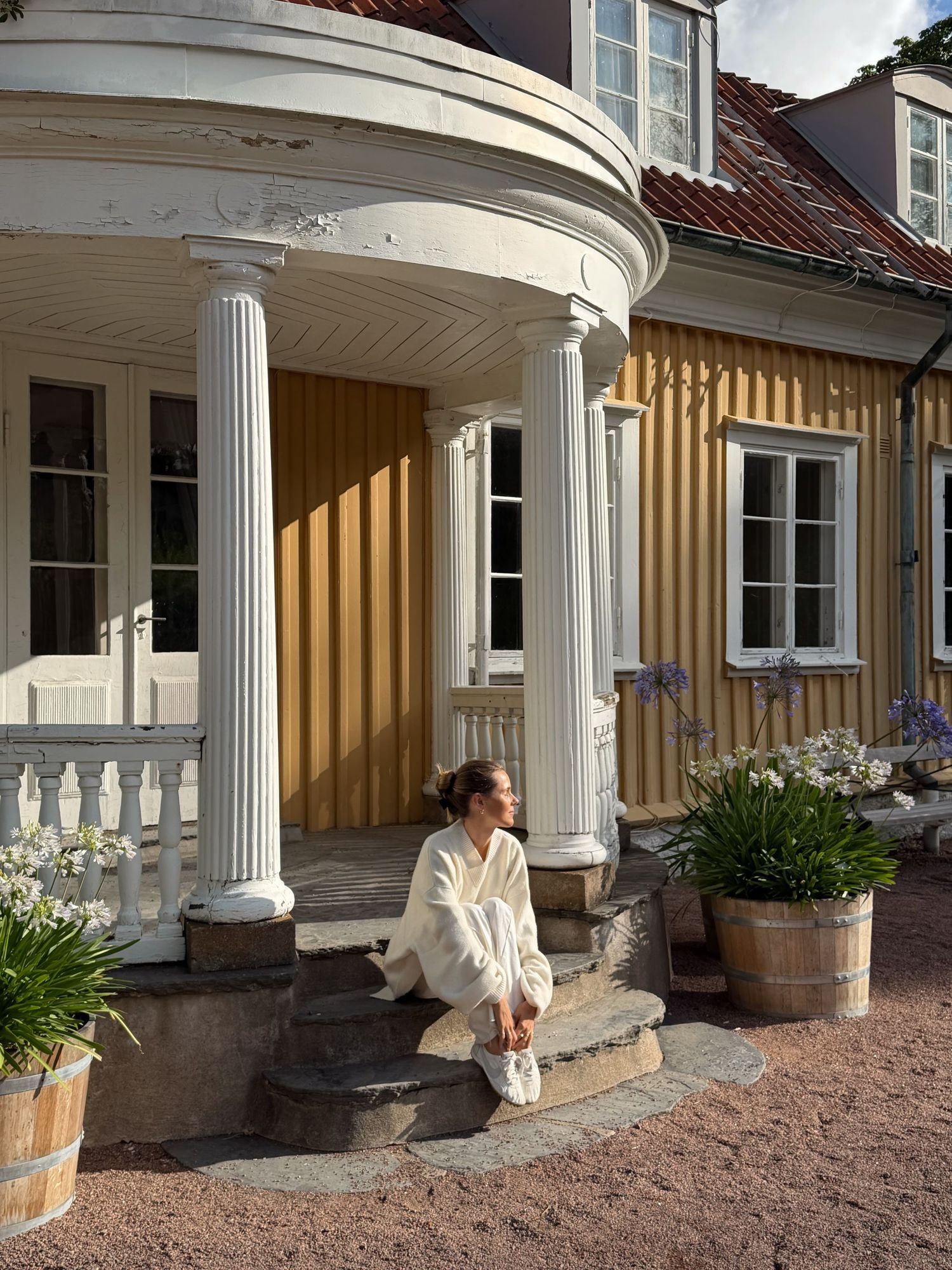 Woman in white outfit sitting on steps of a charming yellow Swedish villa with white columns and lush garden.