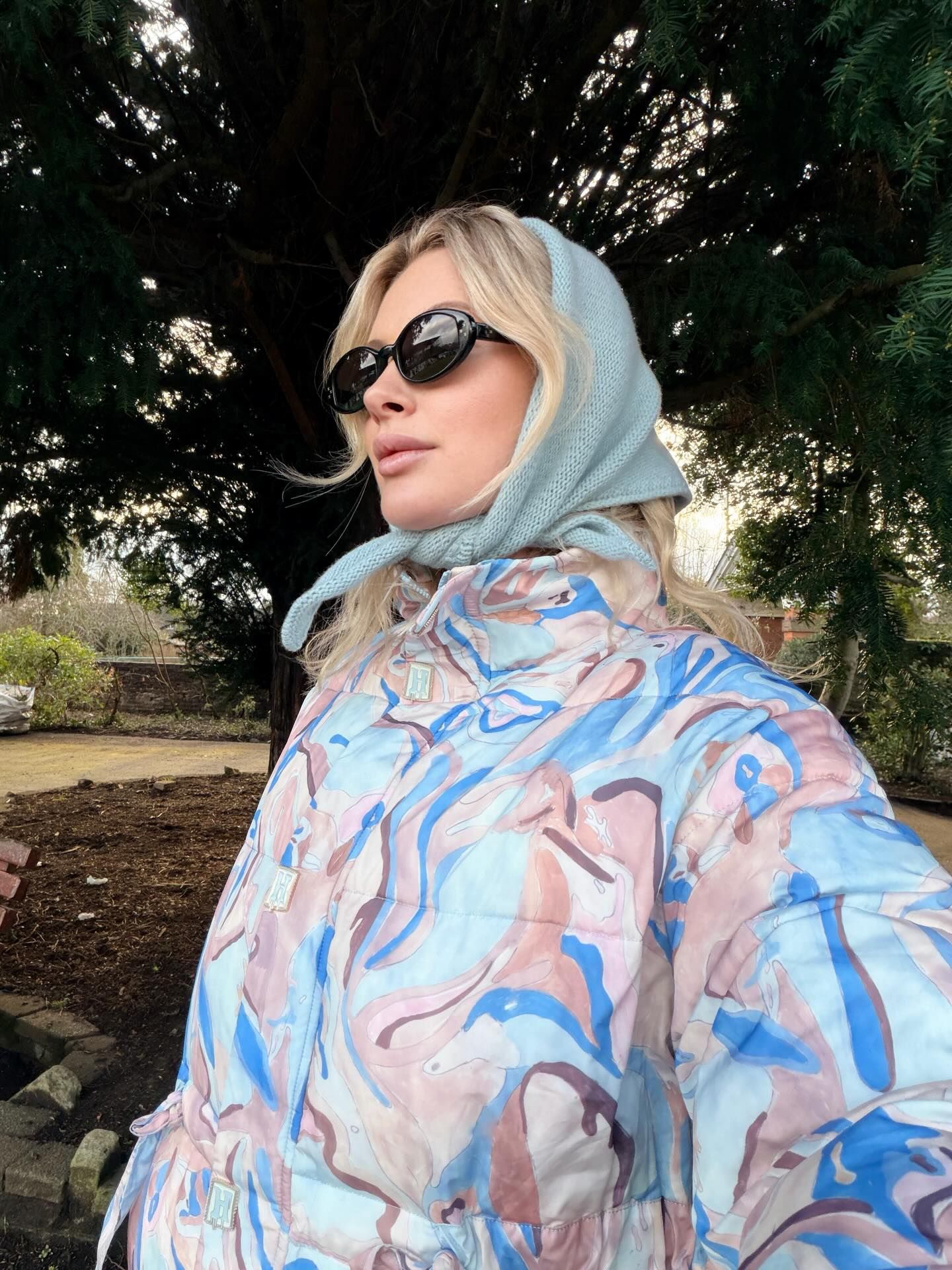 Blonde woman wearing marble print puffer jacket, light blue headscarf, and black oval sunglasses outdoors.