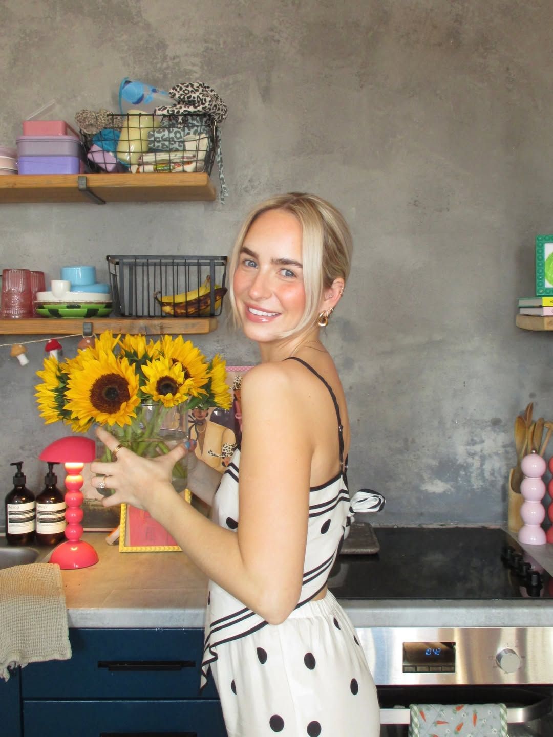 Smiling blonde woman in polka dot dress arranging sunflowers in a colorful, eclectic kitchen with open shelves.