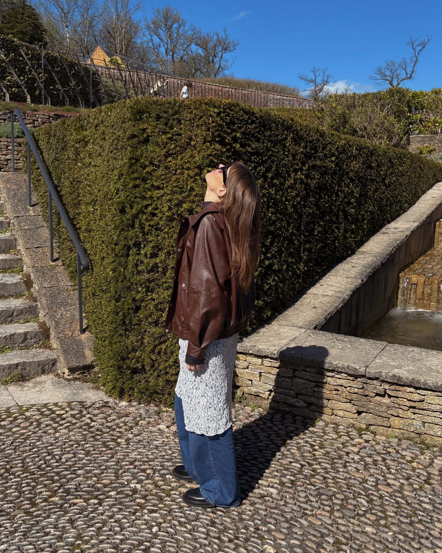 Woman in brown leather jacket and wide-leg jeans standing on cobblestone path beside manicured hedge on sunny day.