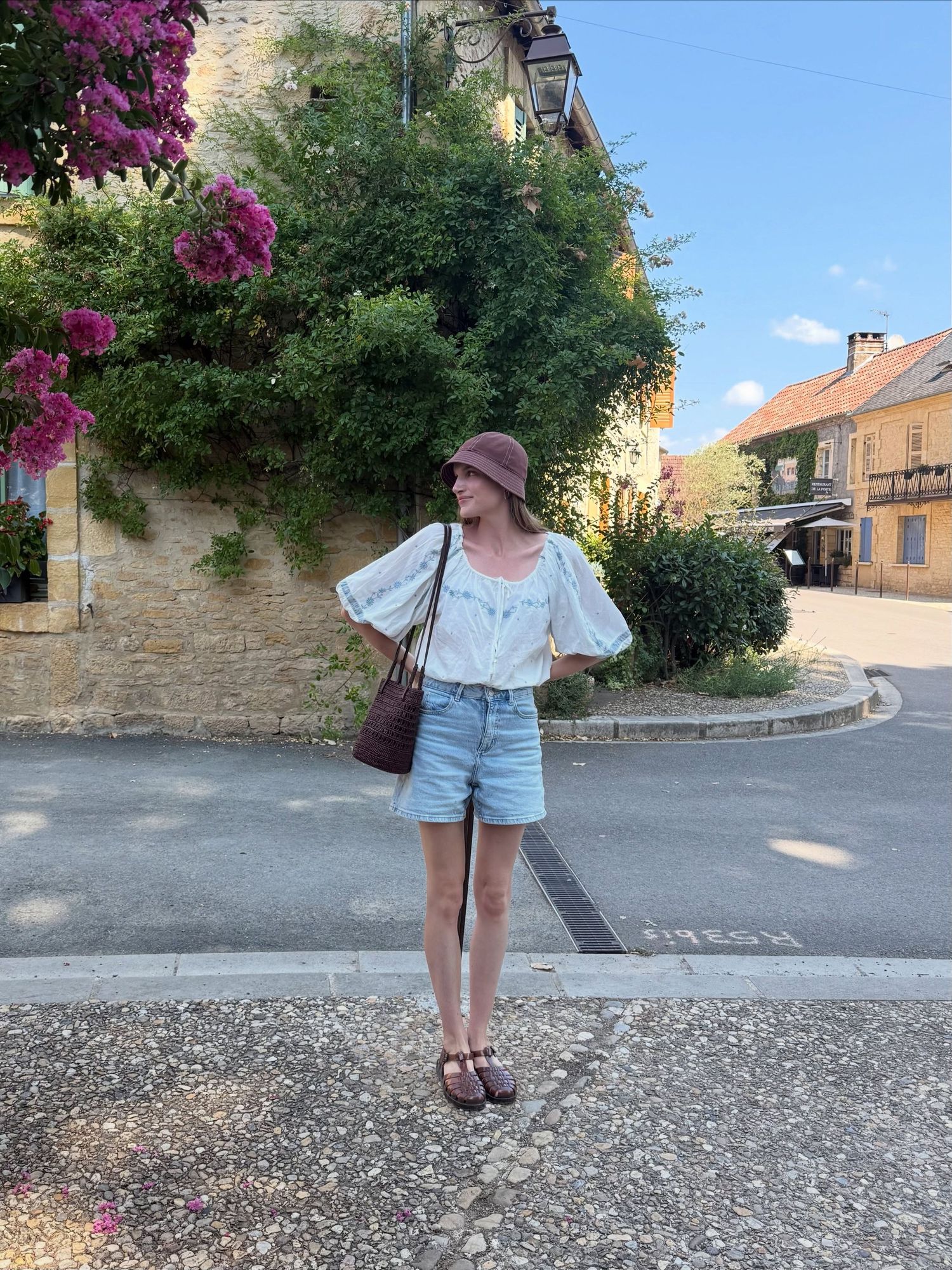 Woman in white blouse, denim shorts, and burgundy bucket hat poses on a sunny European village street.
