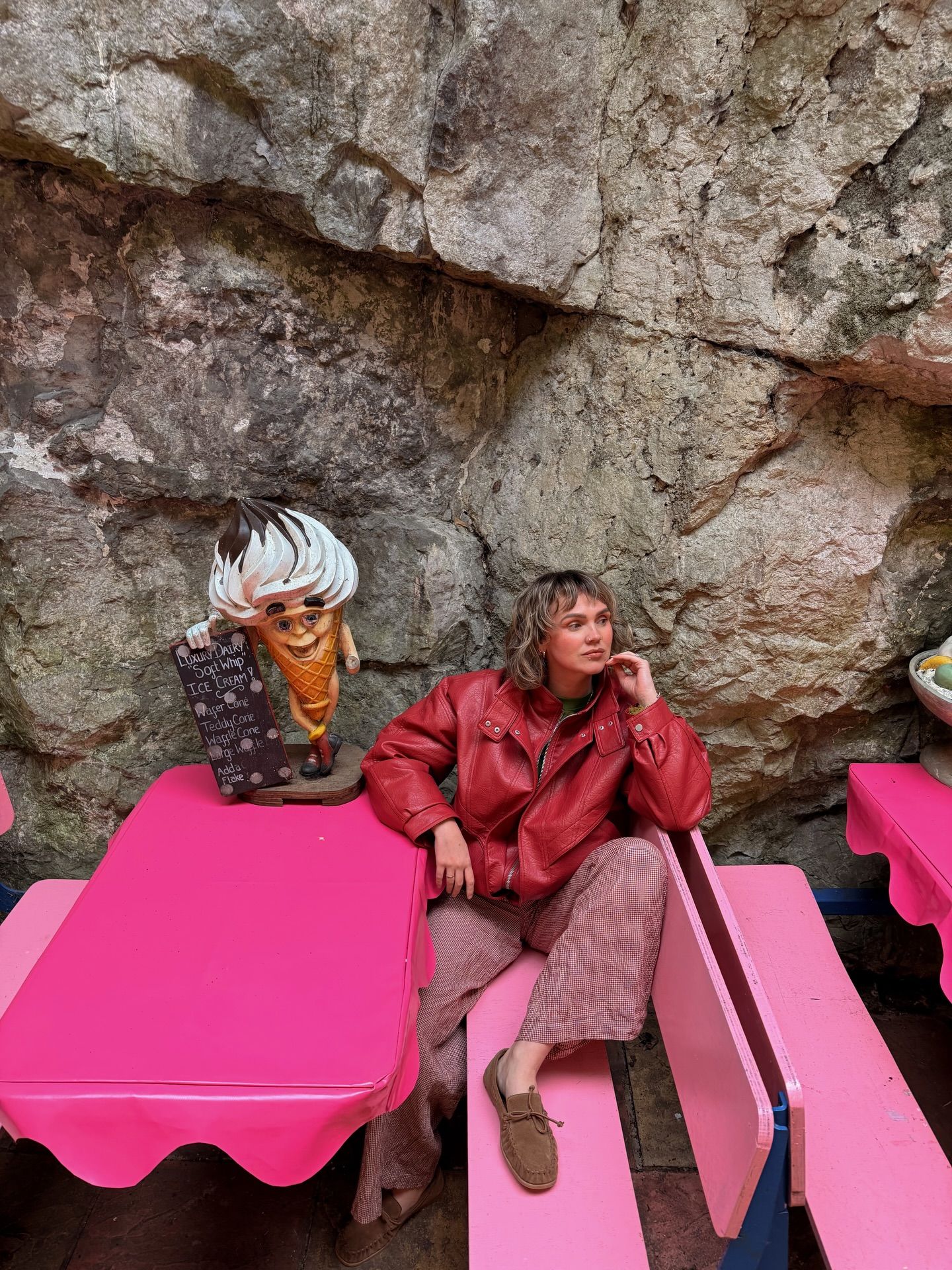 Woman in red leather jacket sits at pink picnic table beside a quirky figurine, against a rocky cave wall backdrop.