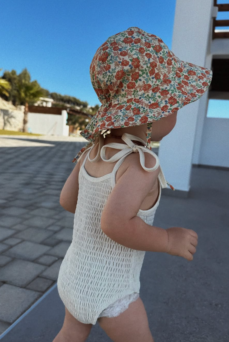 Baby in white smocked swimsuit and floral sunhat playing on a sunny patio.