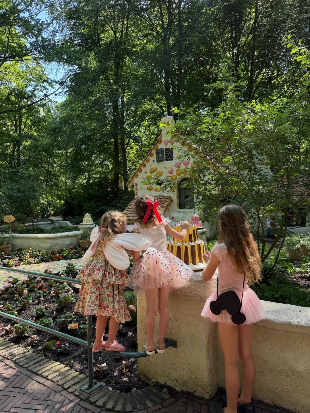 Three girls in fairy costumes explore a storybook gingerbread house display in a lush garden setting.