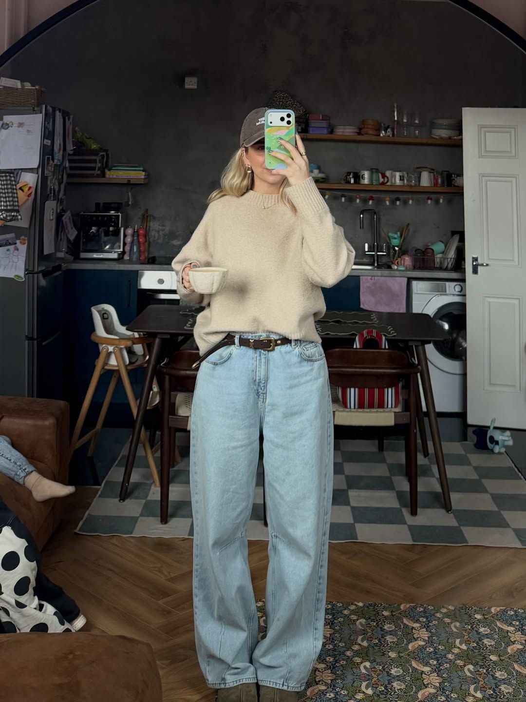 Woman in beige knit sweater and wide-leg light wash jeans taking mirror selfie in stylish kitchen.