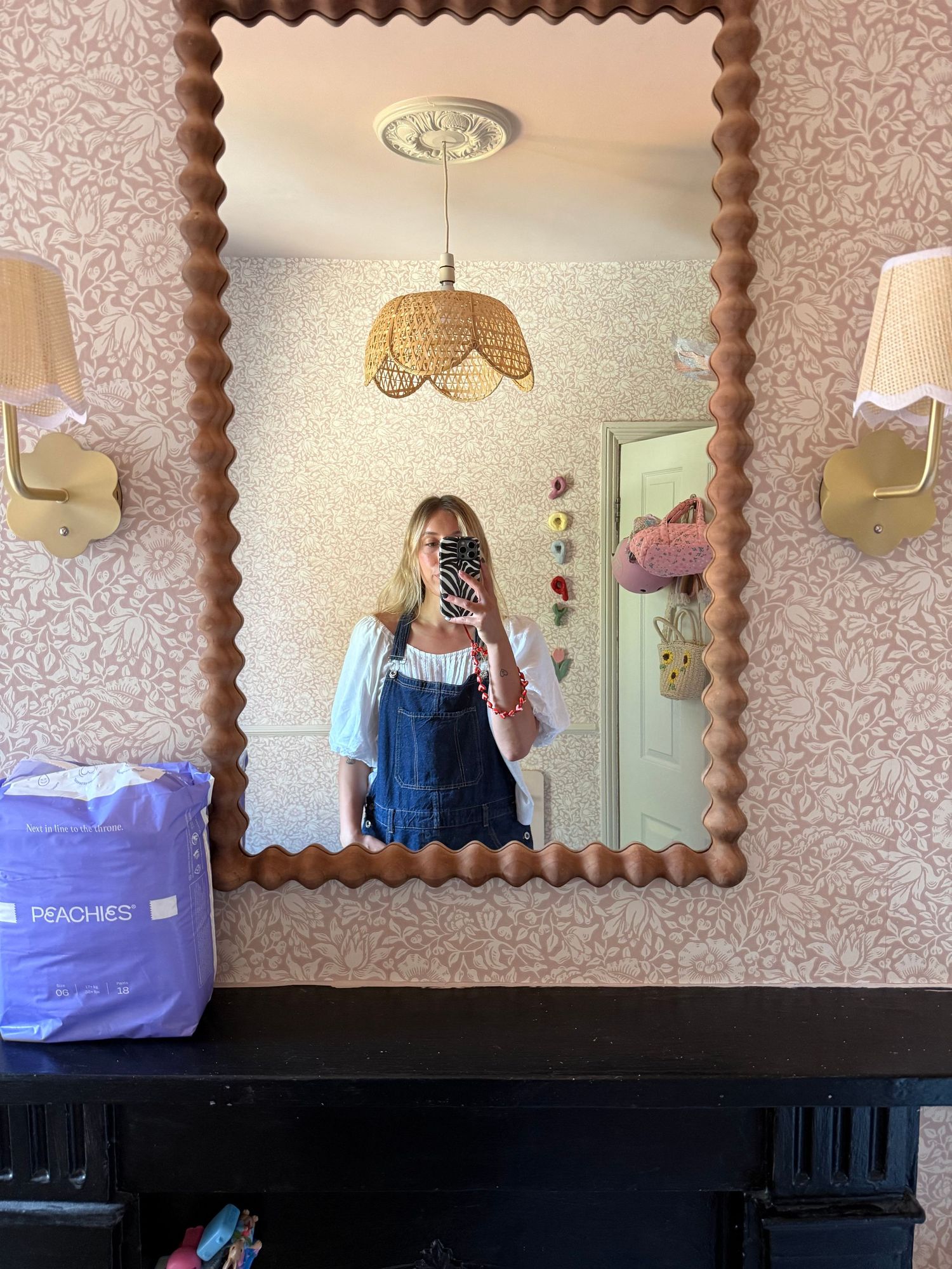 Woman in denim overalls takes mirror selfie in stylish room with scalloped wooden mirror and floral wallpaper.