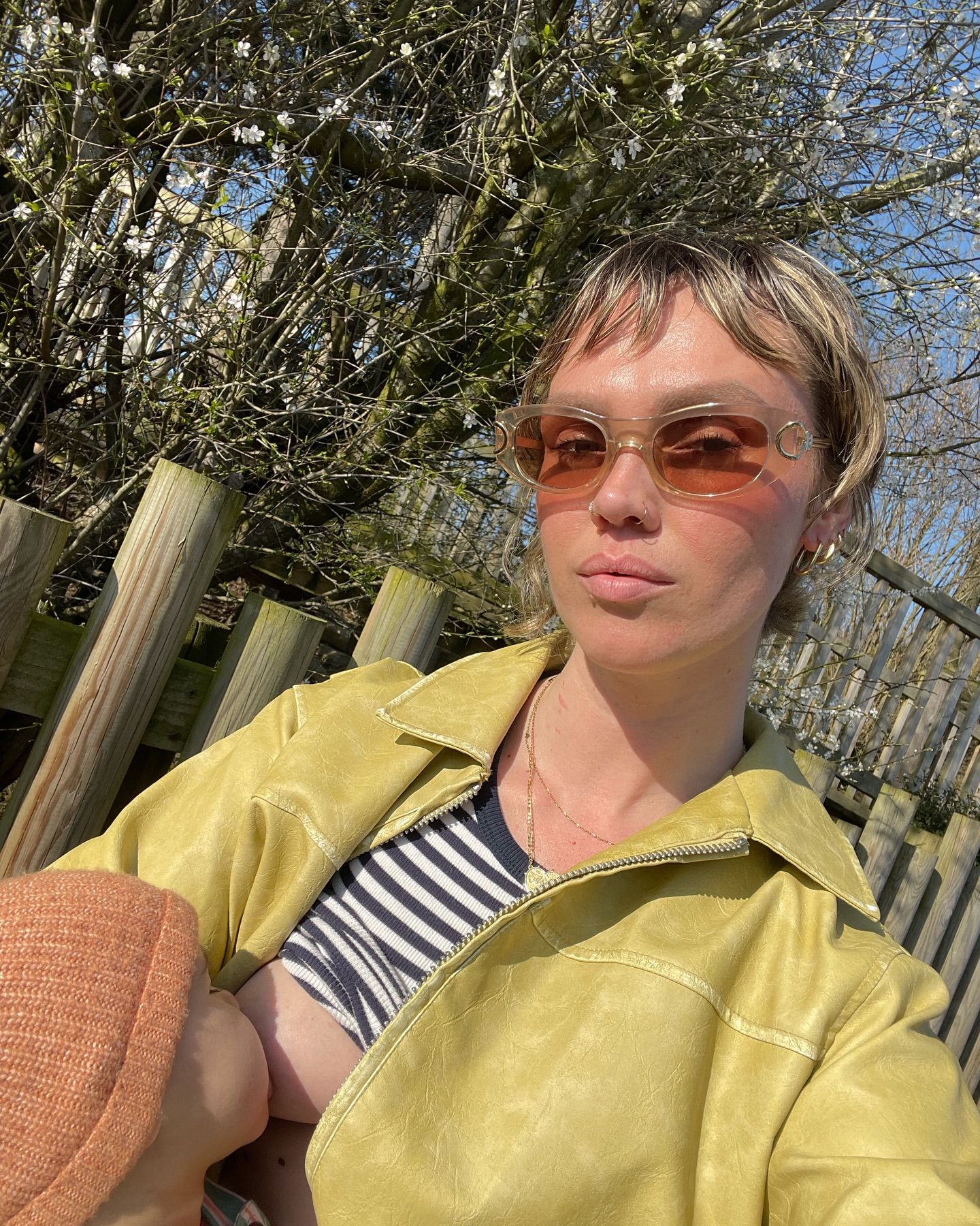 Person wearing yellow leather jacket and pink tinted sunglasses sitting outdoors near a wooden fence and blooming trees.