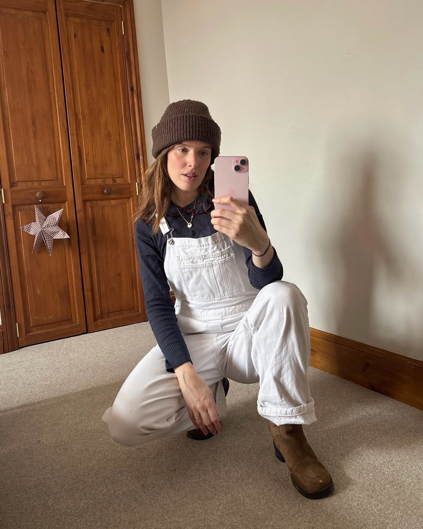 Woman in white overalls, navy top, brown bucket hat and boots takes mirror selfie while crouching on floor.