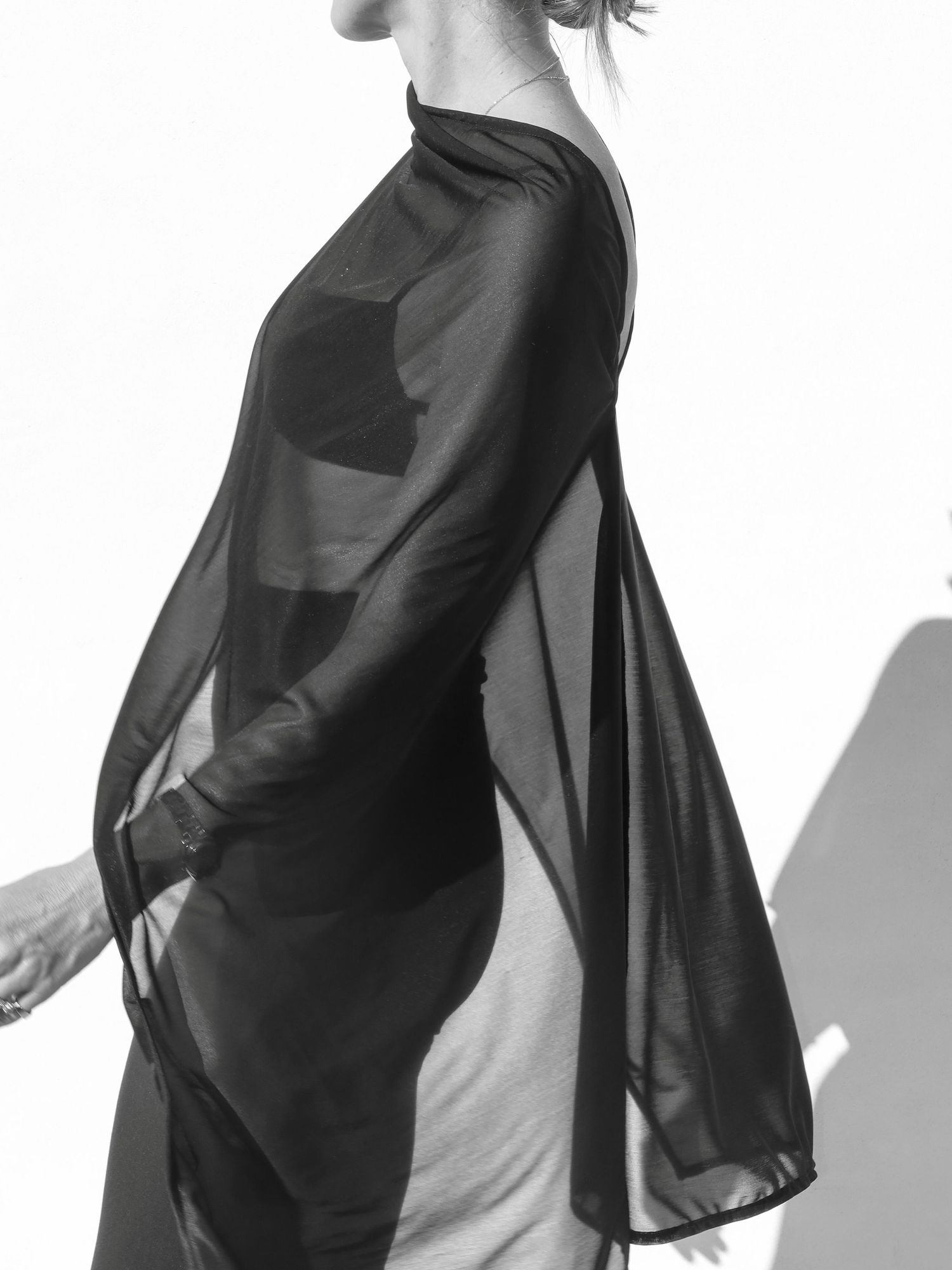 Black and white photo of a woman wearing a sheer black draped top with long sleeves against a white background.