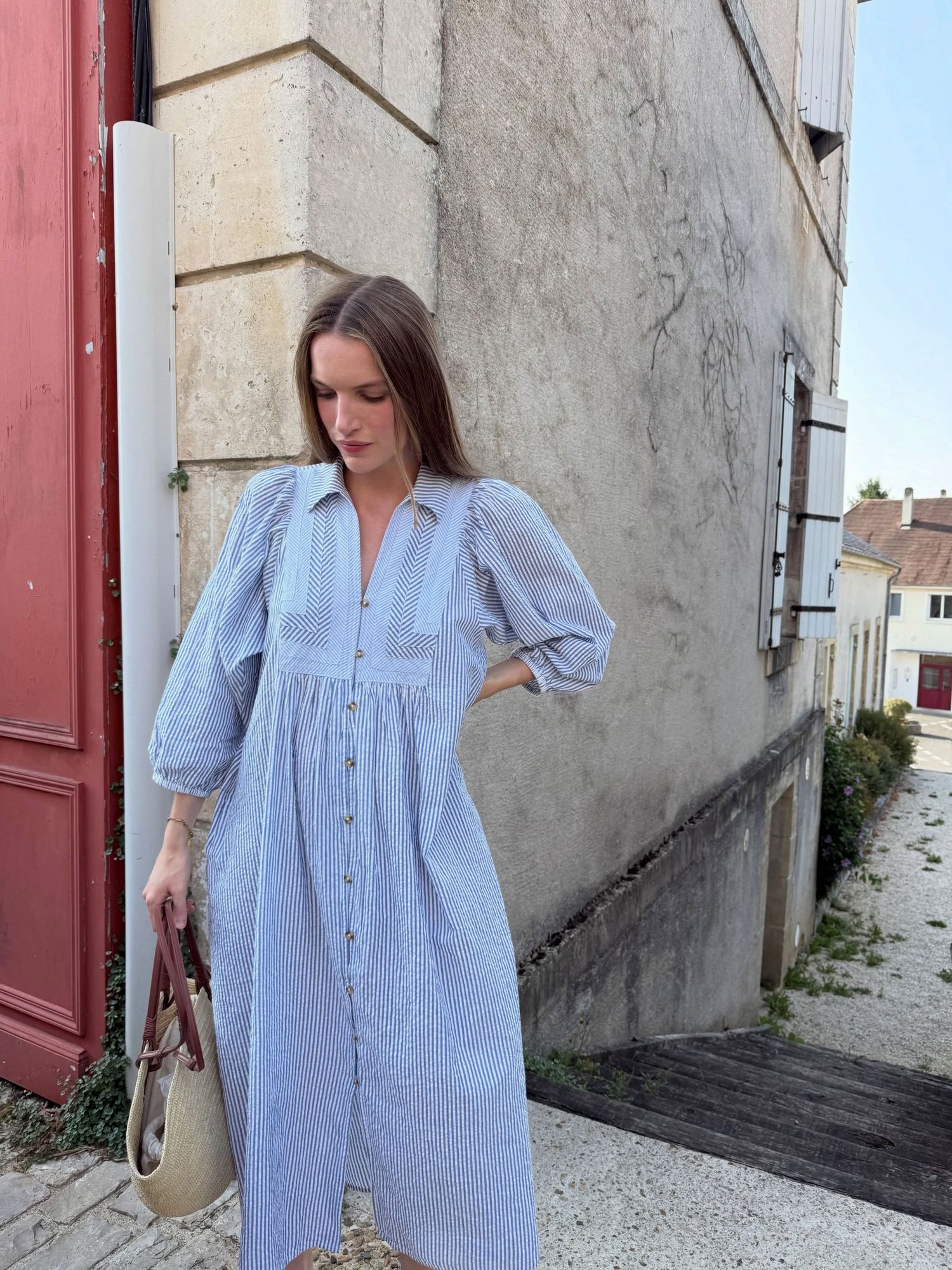 Woman in blue striped midi dress with puff sleeves and buttons, standing by stone building with red door.
