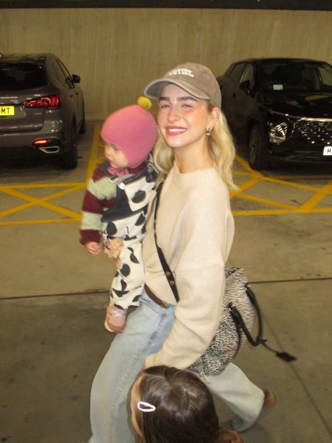 Smiling blonde woman in cream sweatshirt and cap holding a baby in a parking garage.