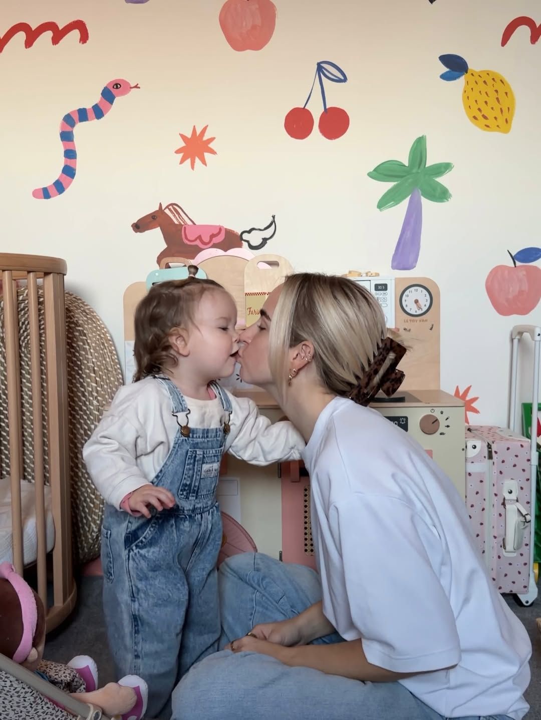A woman kisses a toddler girl in denim overalls in a colorful playroom with fruit and animal wall decals.