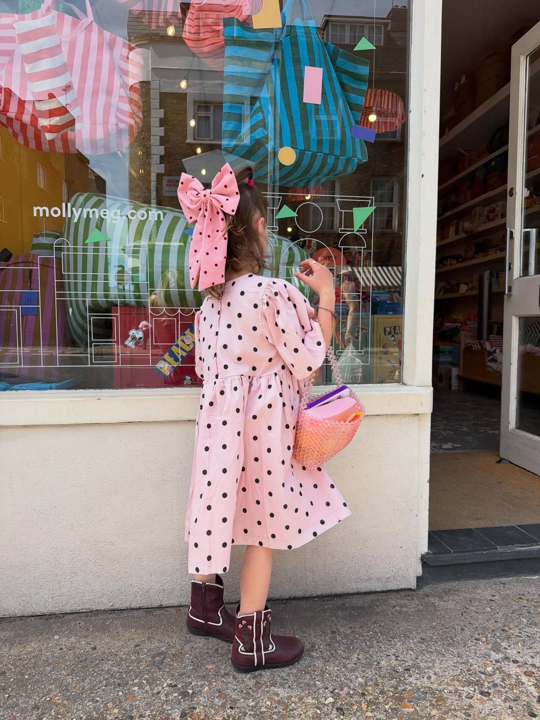 A young girl in a pink polka dot dress and bow stands outside Molly Meg store wearing brown boots holding a colorful bag.
