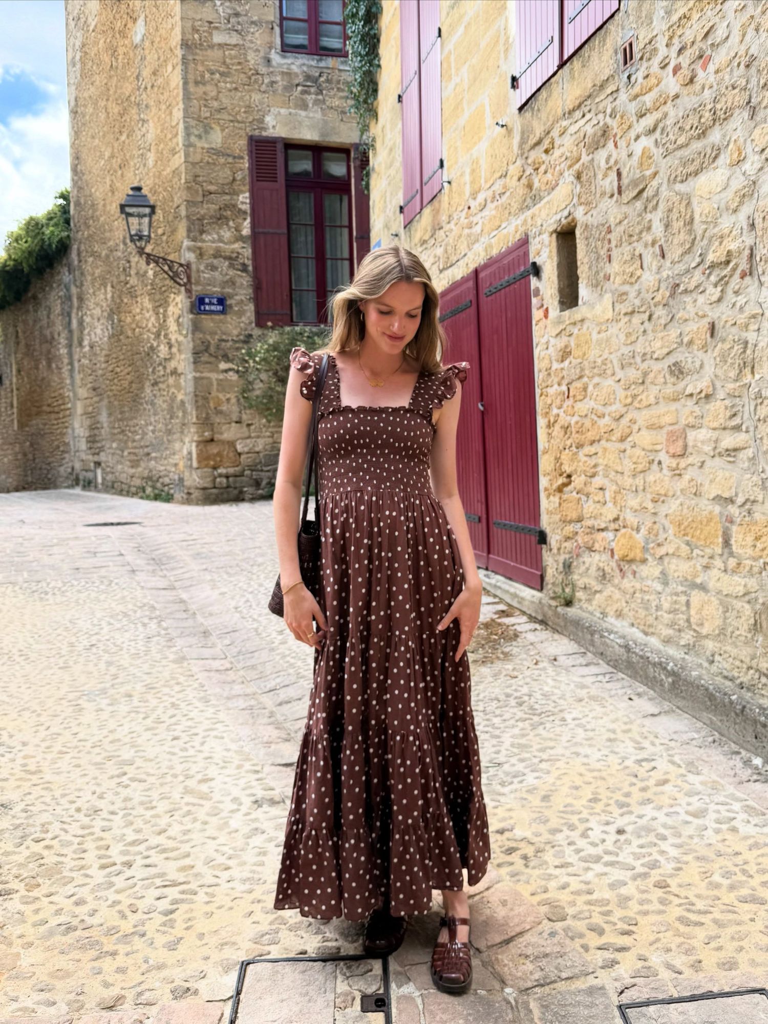 Woman in brown polka dot maxi dress standing on cobblestone street beside stone building with red shutters.