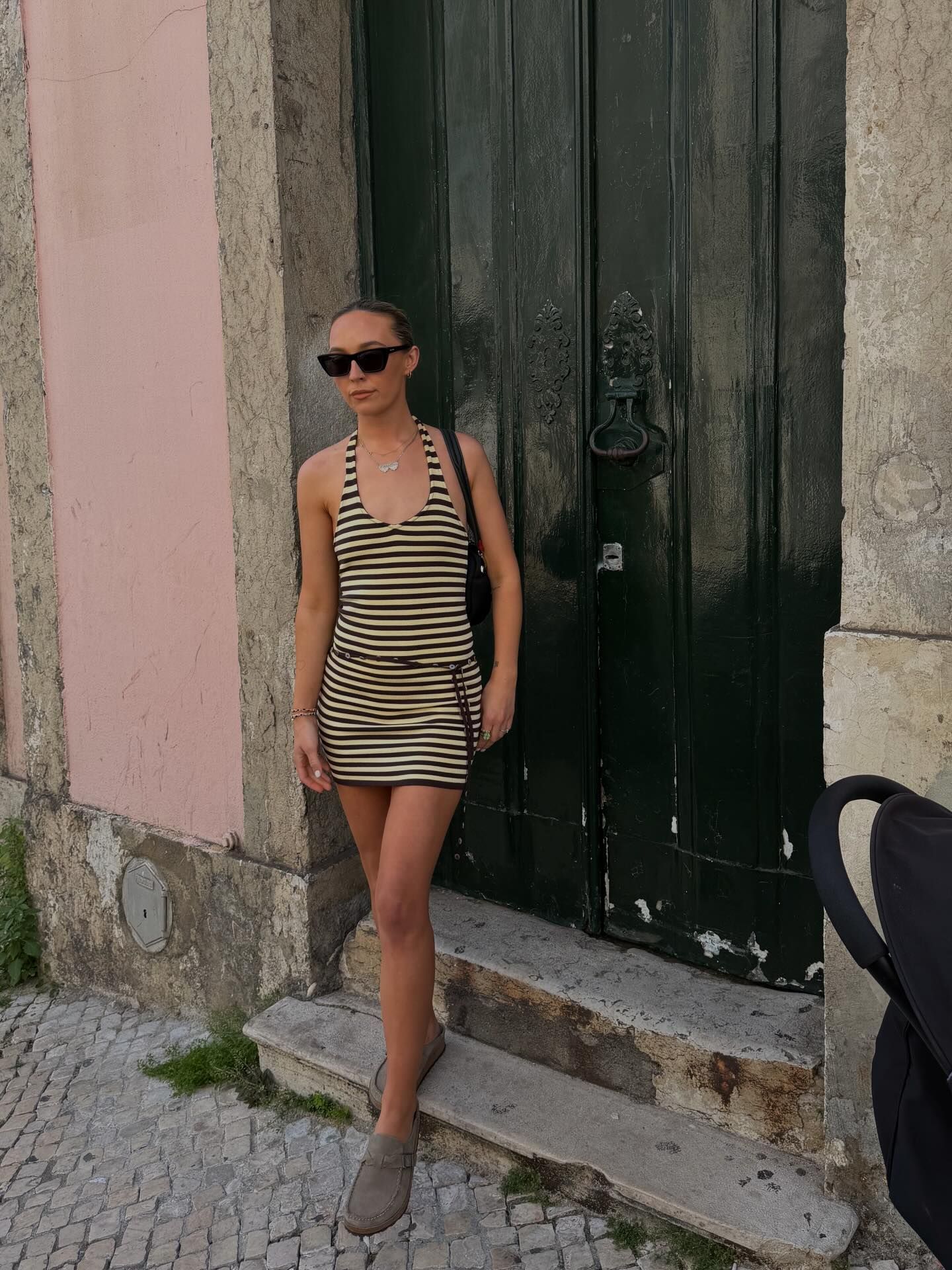 Woman in striped mini dress and sunglasses posing by a vintage black door on cobblestone steps.