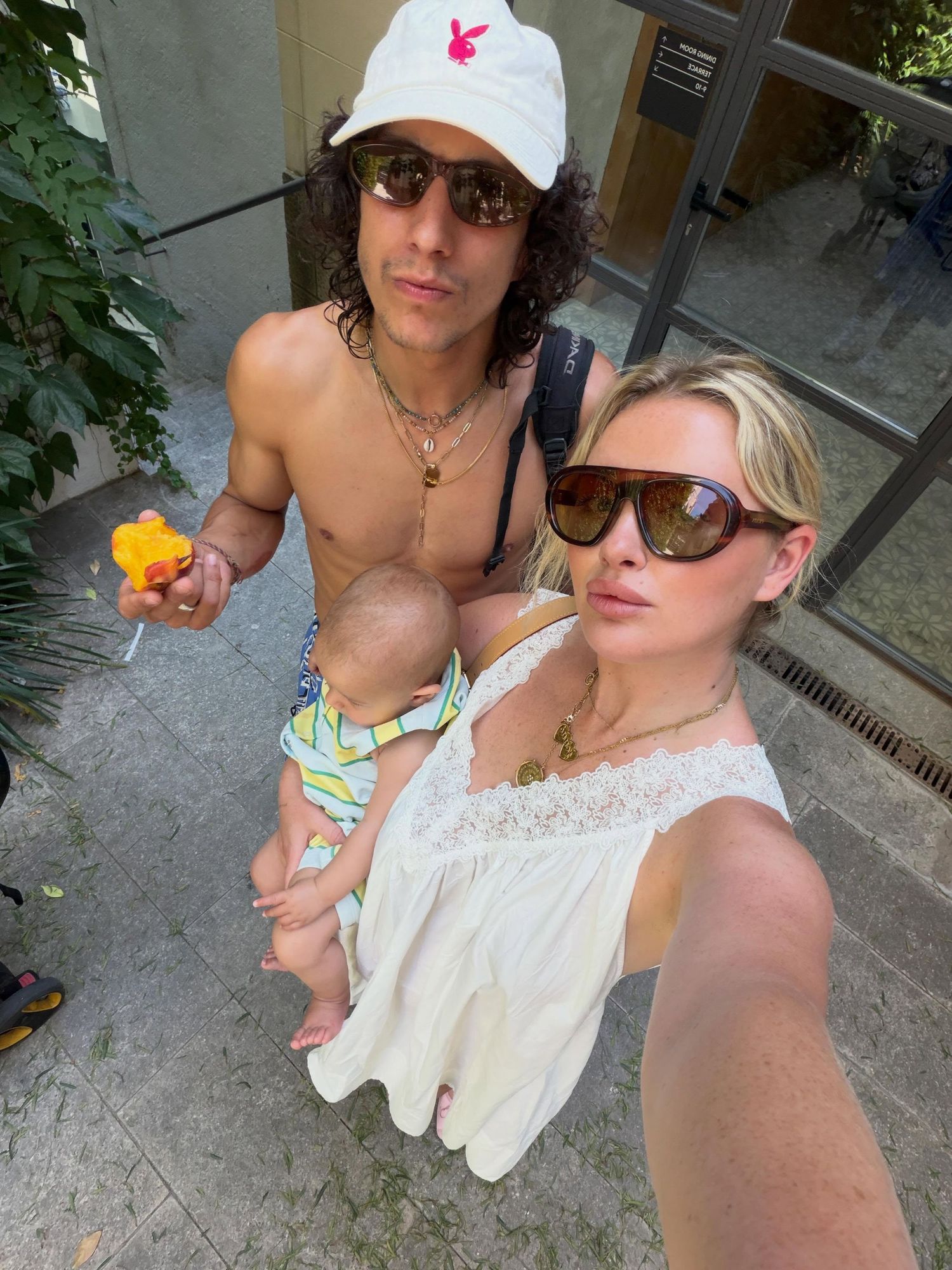 Young couple with baby taking a selfie outdoors, man in white cap holding candy, woman in sunglasses wearing white dress.