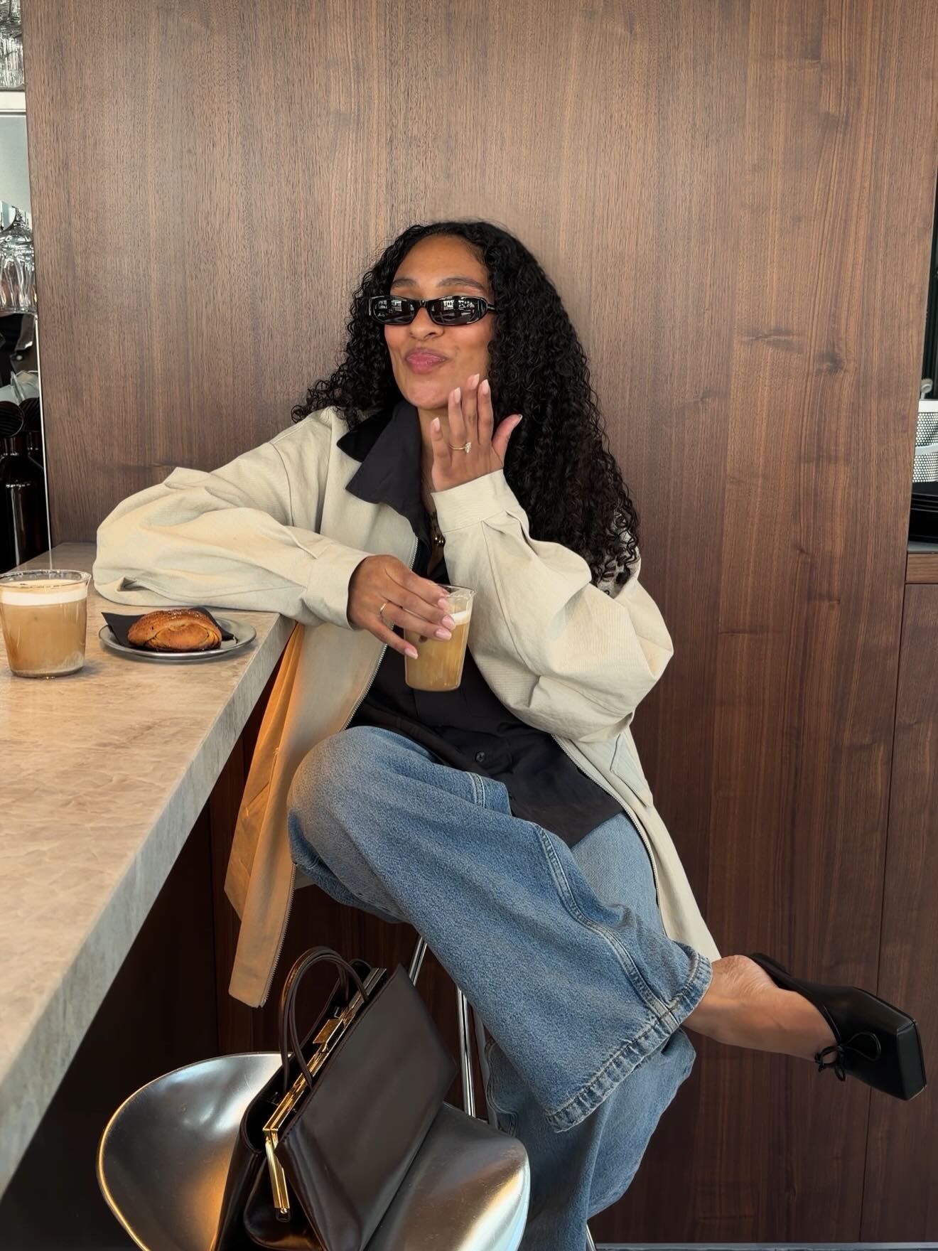Woman in cream jacket, jeans, sunglasses blowing a kiss at a cafe counter holding iced coffee.