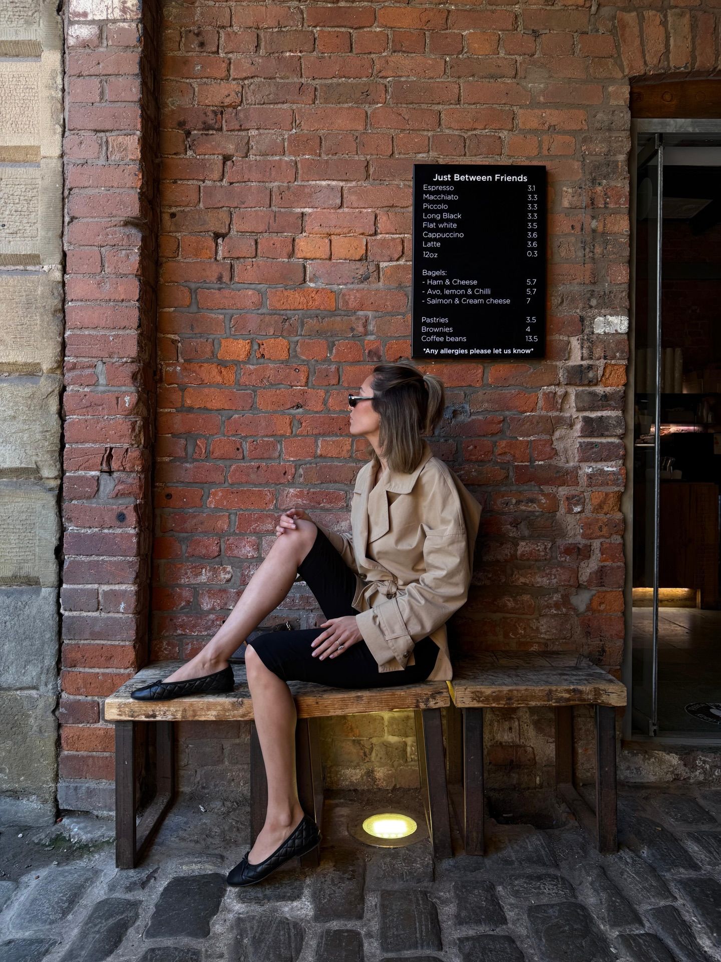 Woman in beige trench coat sitting on wooden bench outside brick wall cafe with menu board.
