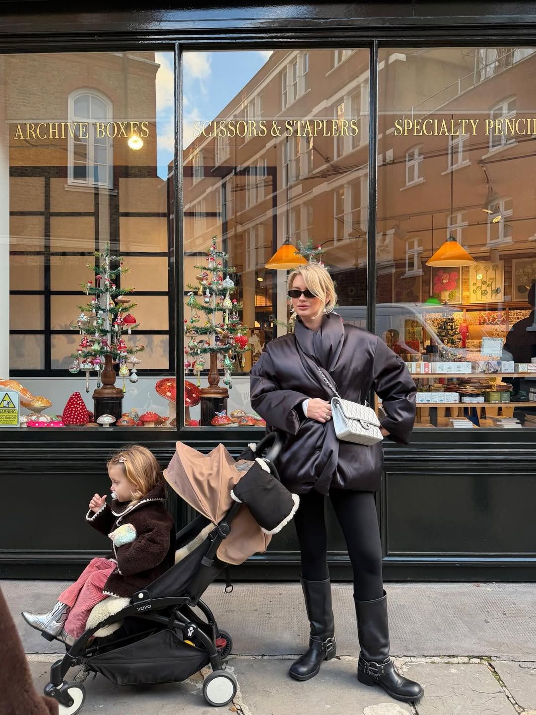Woman in black oversized jacket with stroller outside a stationery shop decorated for Christmas.